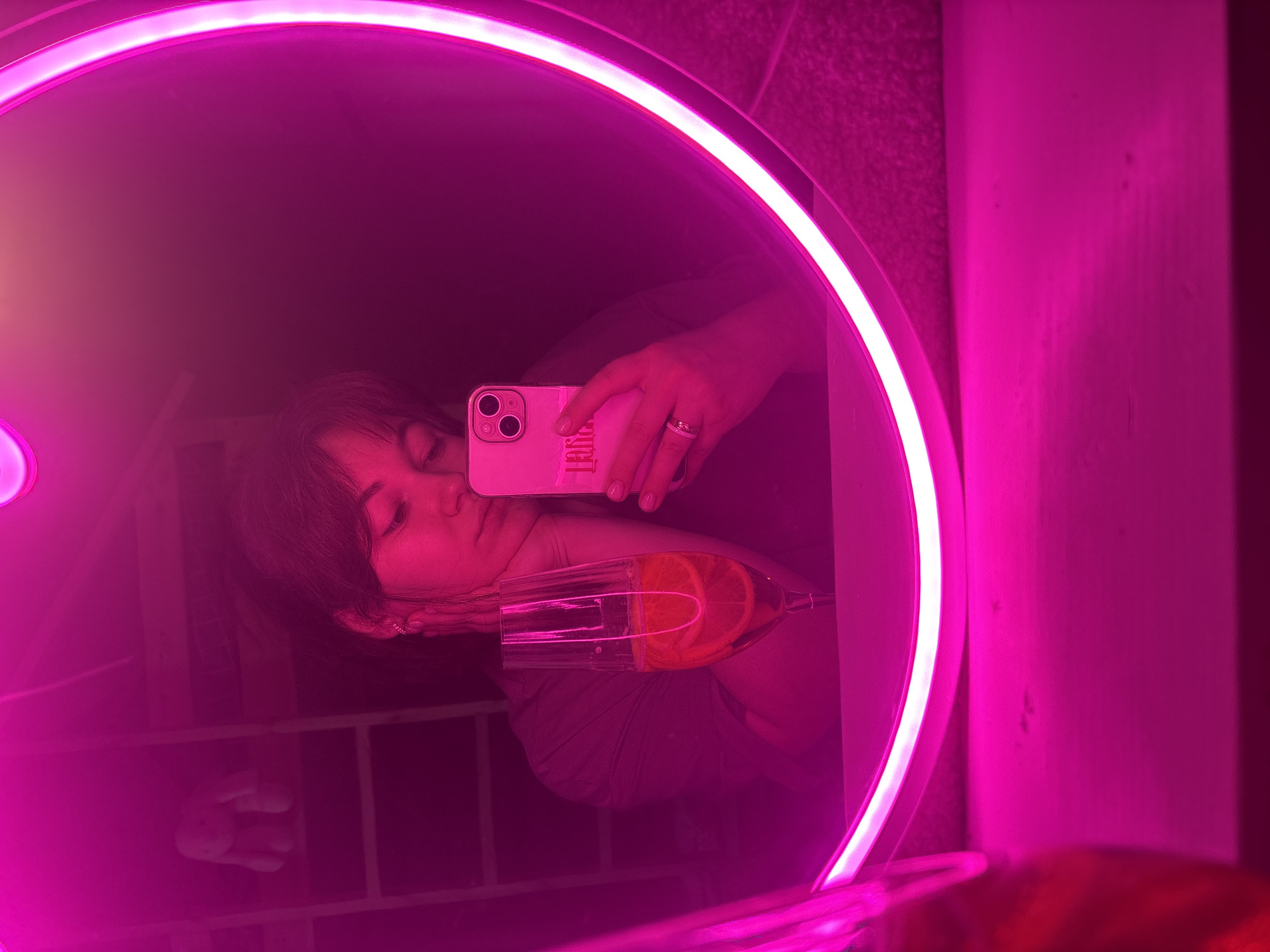 A woman is taking a selfie in a mirror illuminated by a pink neon light ring. She is holding a pink iPhone in her right hand, and a champagne flute with orange slices in her left. She is wearing a ring on her left ring finger and earrings. The background is dark and appears to be an indoor setting. There is no visible indication of the location being in Chaikovsky, Russia.
