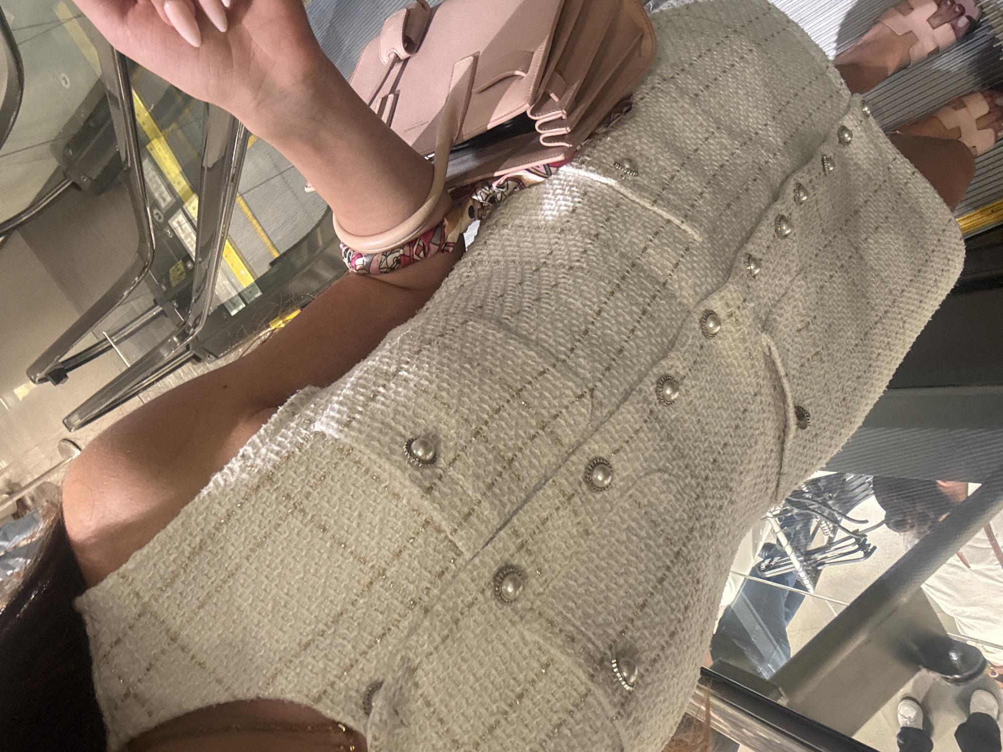 A person stands on an escalator in a brightly lit indoor environment. They are wearing a sleeveless white tweed dress adorned with pearl buttons and gold threads. A pale pink handbag is held in their right hand, and a colorful patterned bracelet is worn on their left wrist. The person's left arm is visible from the shoulder down to the wrist. The background shows the metallic structure and steps of the escalator. The location is Aventura, United States of America.