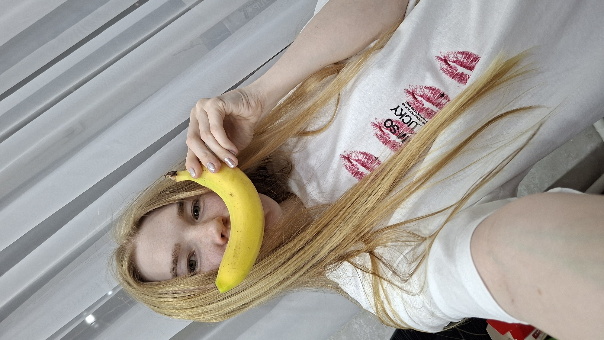 A woman with fair skin and long blonde hair is lying down and holding a yellow banana in front of her face, obscuring her nose and mouth. She is wearing a white t-shirt with lipstick kiss prints and the word "LUCKY" printed on it. The background appears to be a white curtain or draped fabric. The location is the city of Chaikovsky, Russia.