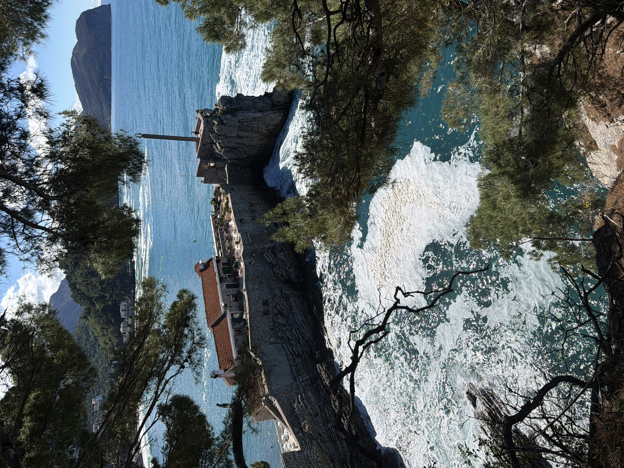 A rotated, eye-level photograph reveals the coastal landscape of Petrovac centar, Montenegro. The scene is framed by tree branches in the foreground. A stone structure with a red-tiled roof, likely a building or part of a historical site, extends out onto a rocky promontory. The Adriatic Sea surrounds the structure, with white foamy waves crashing against the rocks. Further out, the sea appears calm and blue, contrasting with the turbulent waters closer to the shore. In the distance, a mountain or large hill rises against the clear sky. The focus is on the juxtaposition of the historical structure, the natural rocky coast, and the expanse of the sea.