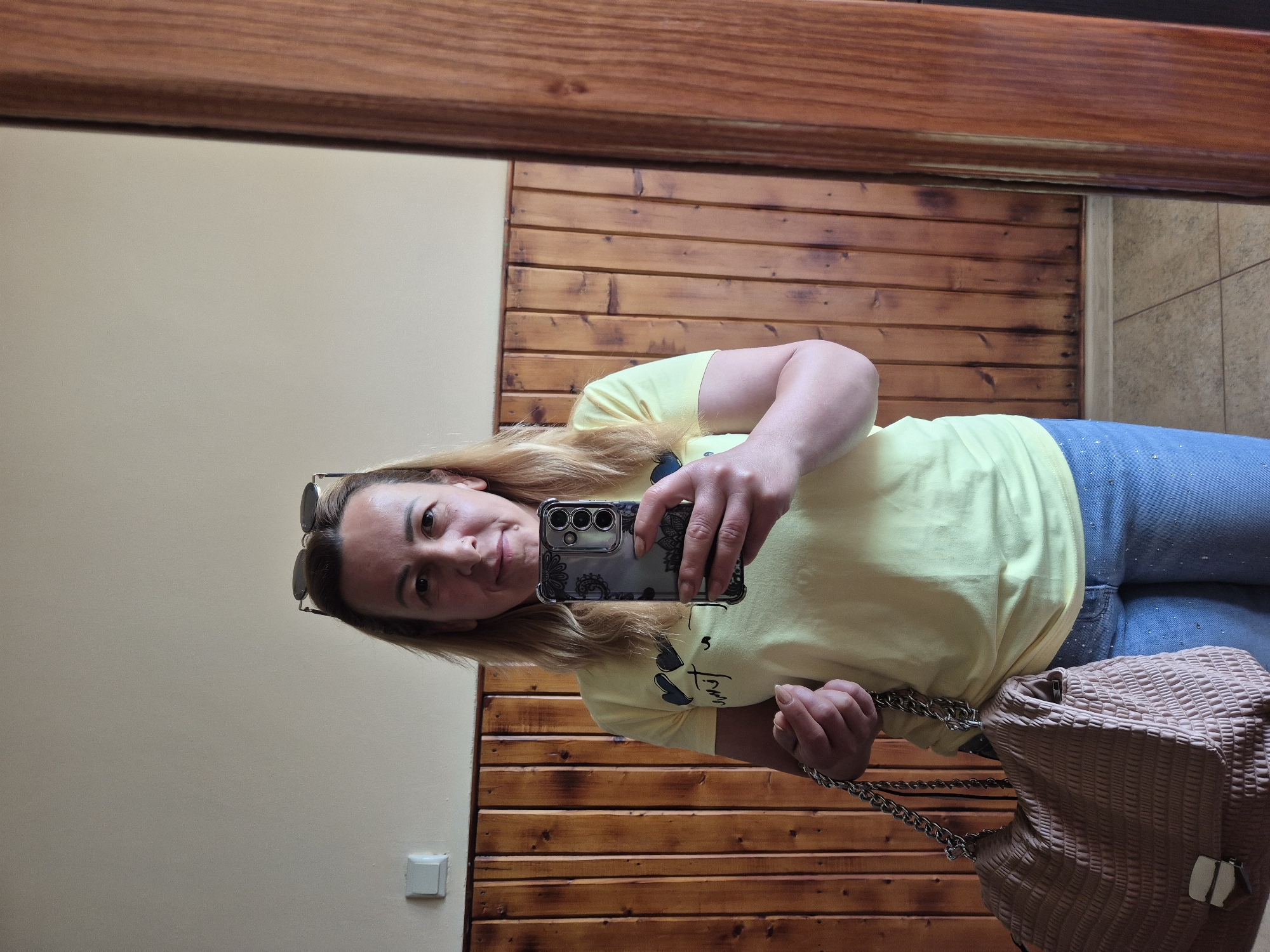A woman with shoulder-length blonde hair, wearing a light yellow t-shirt and blue jeans, takes a selfie in a mirror. She has sunglasses perched on her head and is holding a smartphone in her right hand, which displays a floral pattern. A light brown, textured handbag with a chain strap hangs from her left hand. The background consists of a wall paneled with vertical wooden planks. To the left of the woman, a white electrical outlet is visible on the wall. The floor appears to be tiled. A portion of a wooden doorframe is visible on the right side of the mirror.