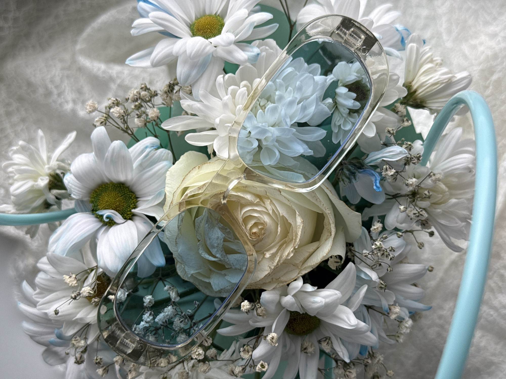 A close-up shot shows a bouquet of white flowers, including daisies and a rose. Overlapping the flowers are a pair of clear eyeglasses. The flowers and glasses are arranged in a light blue container. The background consists of a textured, light-colored surface. There is no indication of the location being Izhevsk, Russia. There are no people present.
