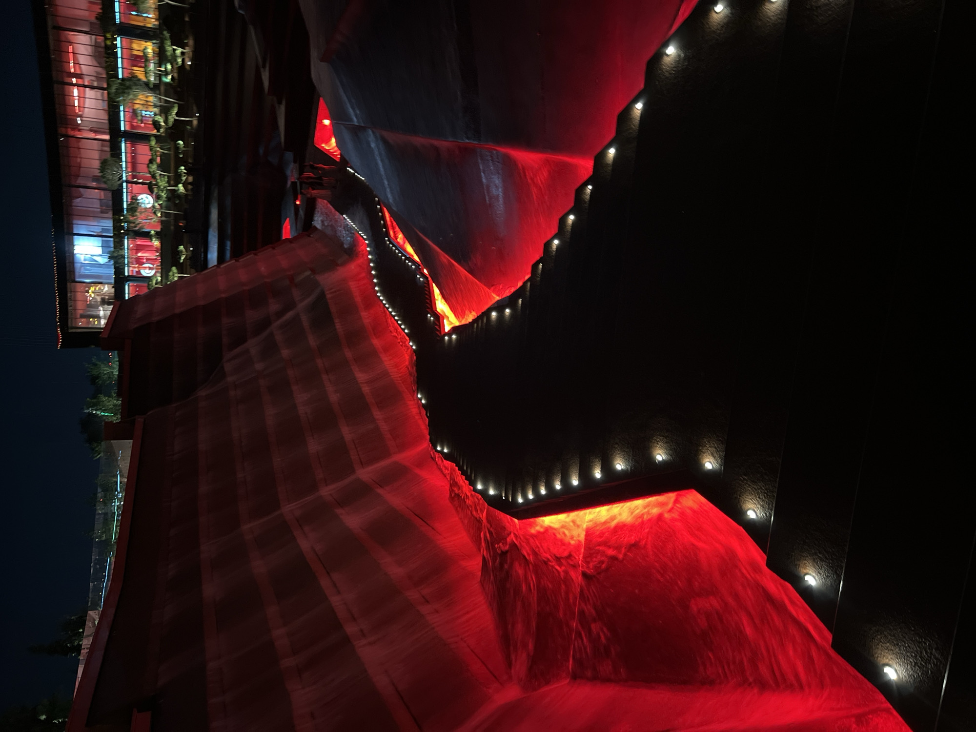 The image captures a nighttime view of a modern architectural design featuring cascading water features illuminated with red light. A dark pathway with small, embedded lights follows the contours of the water channels. In the background, a contemporary building with large glass windows is visible, also lit from within, suggesting it is a commercial or entertainment venue. The overall scene evokes a sense of dramatic ambiance. The location is the "Water Garden" park in Krasnodar, Russia.