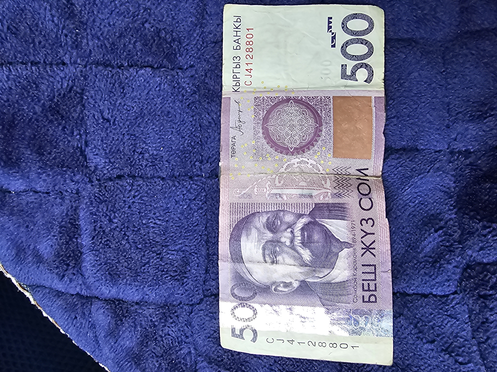 A close-up, top-down shot shows a blue textured blanket with a Kyrgyzstani 500 som banknote laid on top. The banknote is oriented horizontally, with the left side featuring a portrait of an elderly man with a mustache and a cap. To the left of the portrait are the numbers "500" in large font and a serial number "CJ4128801". The right side of the banknote has the text "КЫРГЫЗ БАНКСЫ" and the denomination "500" in large font. The banknote is slightly creased. The image does not contain any visual information about the city of Özgön, Kyrgyzstan.