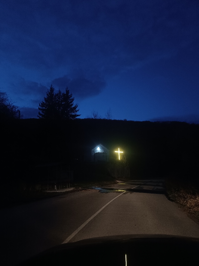 The image depicts a nighttime scene in Oreshets, Bulgaria, captured from a vehicle's interior. A portion of the vehicle's hood is visible at the bottom. An asphalt road with a single white solid line extends into the midground, showing reflective wet patches on its surface. The sky is dark blue, indicating twilight or night, with visible clouds. To the left, a dense silhouette of evergreen trees outlines a distant hillside. To the right, a darker hillside rises. Positioned on this right hillside are two illuminated subjects: a small, white-colored structure, likely a chapel or shrine, illuminated by a distinct white light source above its entrance, and immediately to its right and slightly higher, a large, prominently glowing yellow cross. No individuals are present.