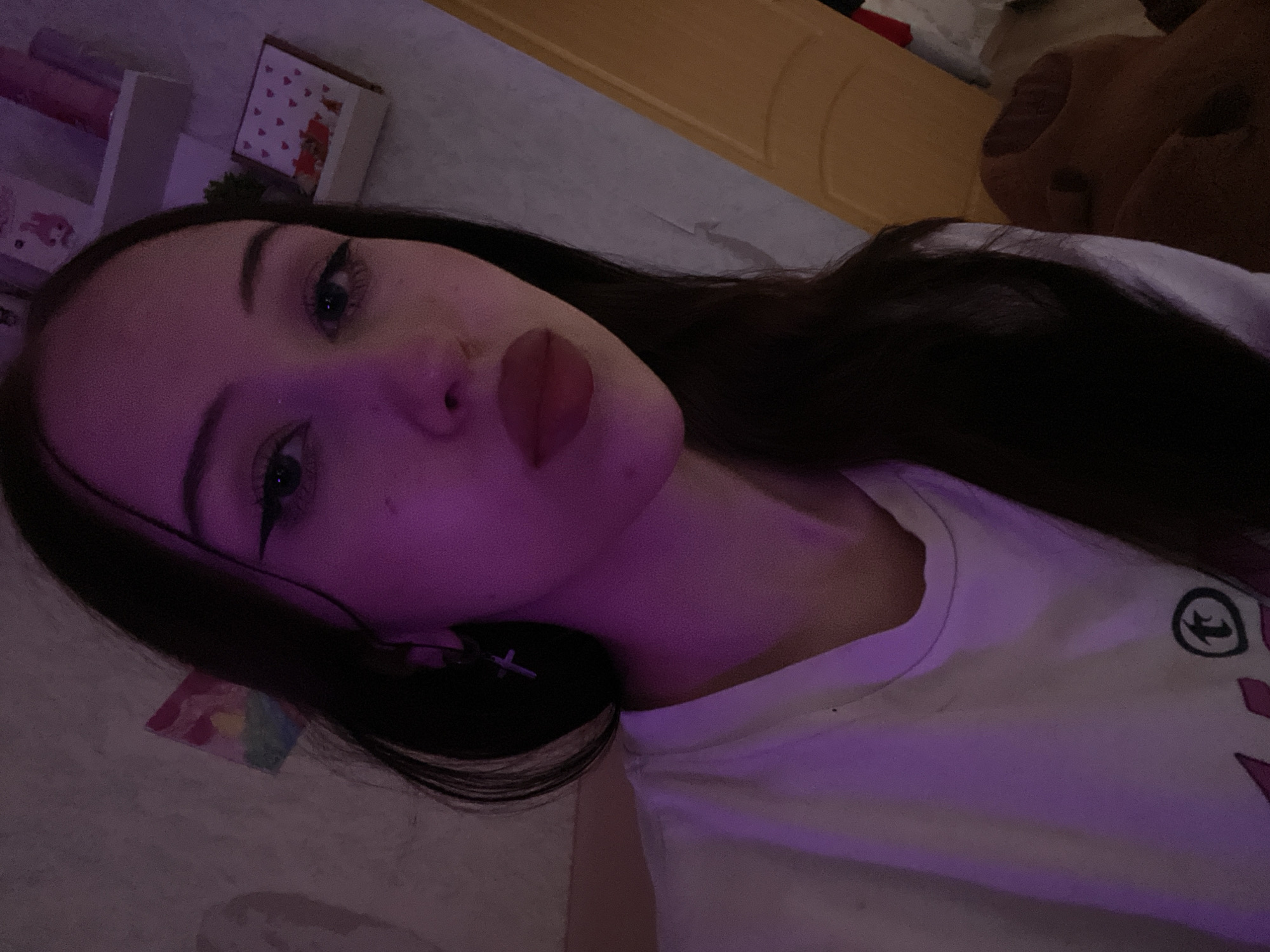 A young woman with dark hair and dark eyes is shown in a close-up, selfie-style shot. She is wearing a white t-shirt and a cross earring. The lighting is dim with a strong purple hue. In the background, there are shelves with various decorative items, including what appears to be a small picture and a box with heart patterns. The overall environment suggests an indoor setting, possibly a bedroom. There is no indication of the specific location within Votkinsk, Russia.