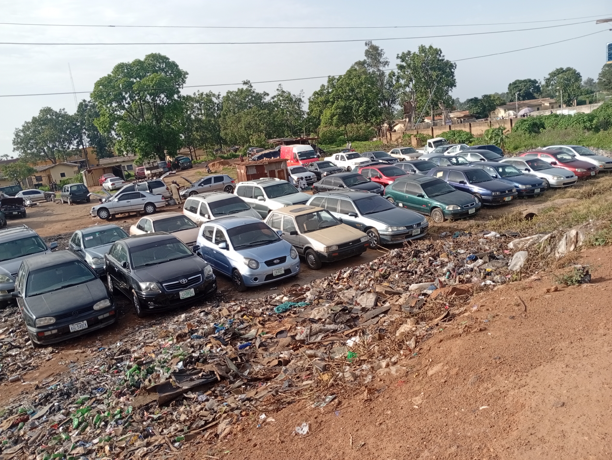 The image depicts an outdoor scene in the city of Rafin Par, Nigeria, characterized by a large accumulation of discarded vehicles and a significant amount of refuse. In the foreground, a steep incline of trash is visible, consisting of various types of waste, including plastic bottles, debris, and organic matter. A row of parked cars is positioned behind this refuse pile.
Among the vehicles, a black Volkswagen Golf with license plate "AGL 54DCK" is prominently displayed. Next to it is a black Toyota Avensis, bearing license plate "LG1 399 AE". Further down the line, a light blue Kia Picanto with license plate "LGN 46778" is parked. Other cars, varying in color and make, are scattered throughout the scene, many appearing to be old or damaged. Some vehicles have their hoods open, suggesting possible repair work or salvage operations.
In the background, a red van is visible, along with more parked cars. Buildings and trees are present in the distance, indicating a semi-urban or rural environment. The overall impression is one of a car junkyard or an area used for discarding old vehicles and waste, likely in a less developed part of the city. The lighting suggests it is daytime, possibly late morning or early afternoon.