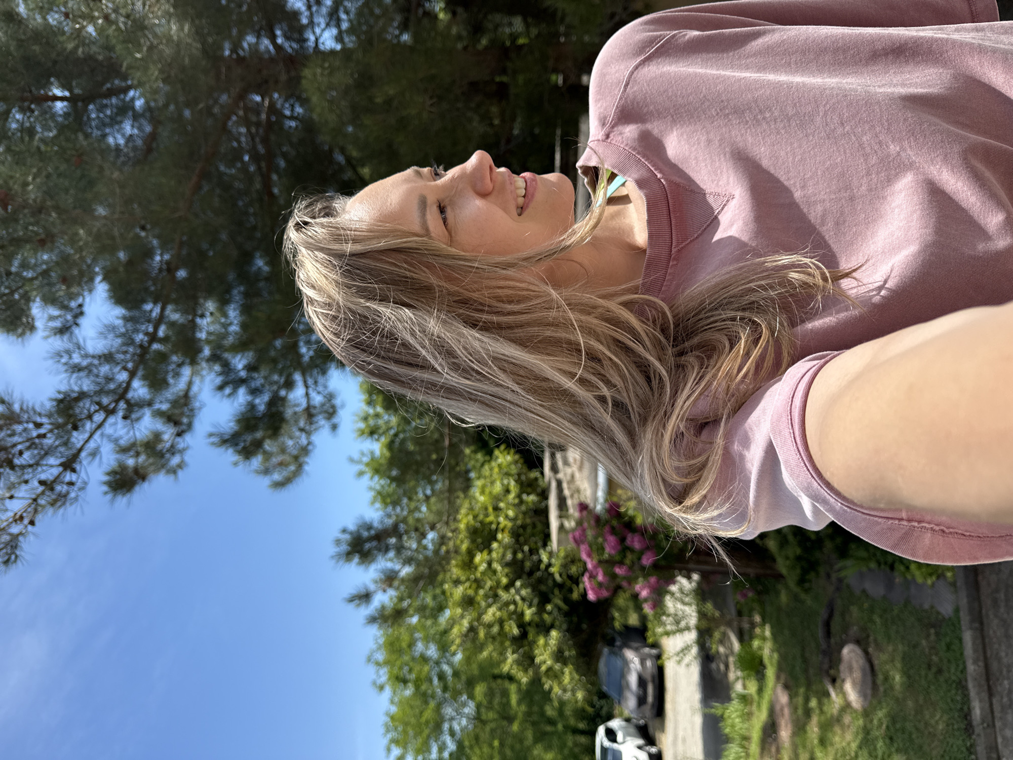 A blonde woman, facing right in profile, smiles as she takes a selfie outdoors. She is wearing a pink sweatshirt. In the background, there are trees, bushes with pink flowers, a blue sky, and cars parked on a street. The location is Nebug, Russia.