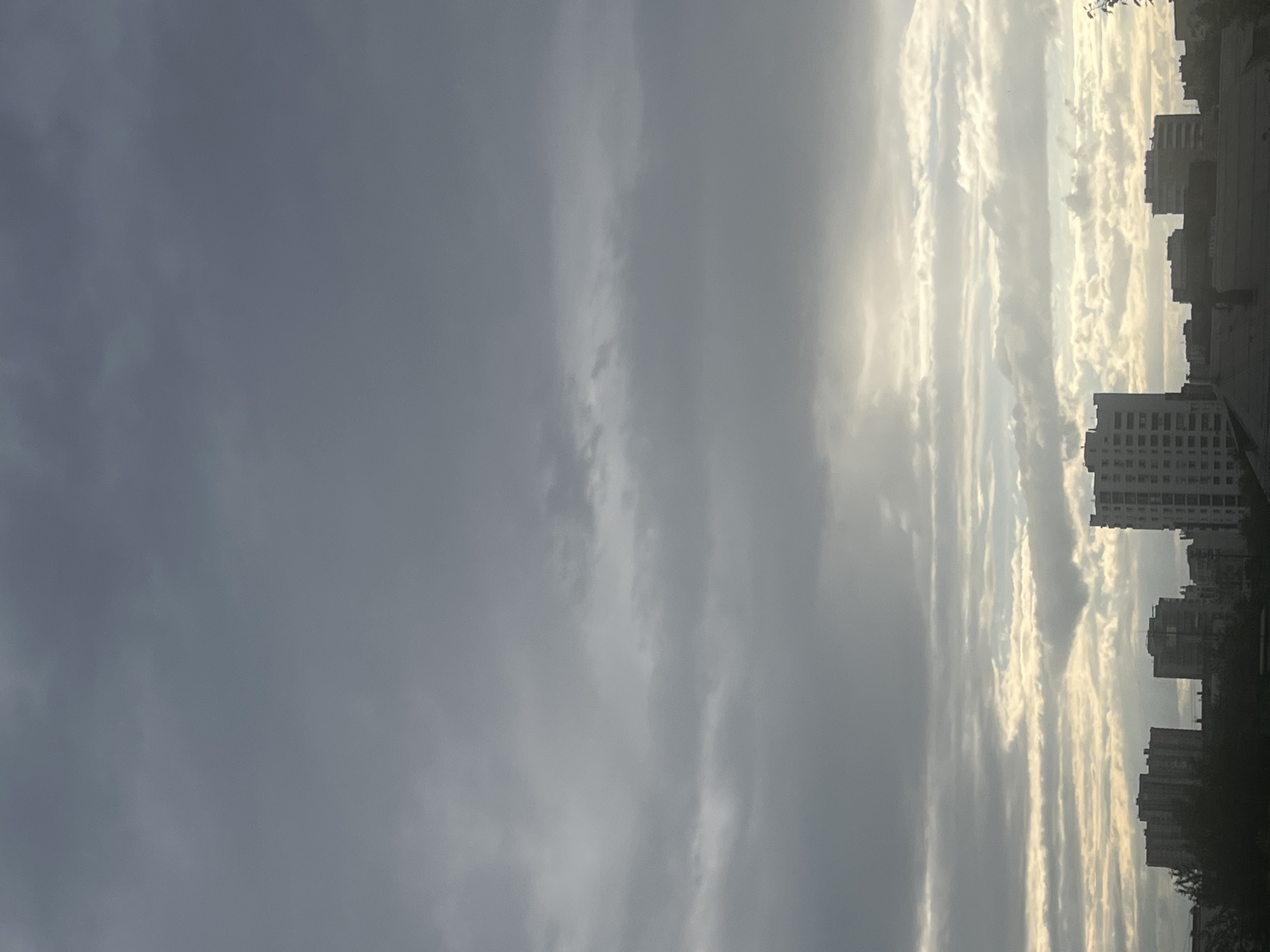This media file captures a dramatic sky over a cityscape. The scene is dominated by a gradient of clouds, transitioning from a dark, stormy gray on the left to bright, sunlit white and yellow clouds on the right. The sun is positioned behind the clouds on the right side of the frame, casting a strong, ethereal glow.
Silhouetted against the bright sky on the right are several multi-story buildings, characteristic of an urban environment. These buildings are dark and indistinct, providing a stark contrast to the illuminated clouds. Trees are also visible in the foreground on the right, their dark forms adding to the silhouette effect.
The time of day appears to be either sunrise or sunset, given the low angle of the sun and the intense backlighting. The weather seems dynamic, with a mix of heavy clouds and bright, sunny patches, suggesting a changeable sky. The overall mood is one of awe and perhaps a touch of melancholy due to the dramatic contrast and the darkness of the buildings. The location is Chelyabinsk, Russia, which contextually aligns with the architectural style of the buildings and the type of sky often seen in this region. No visible text is present in the image.