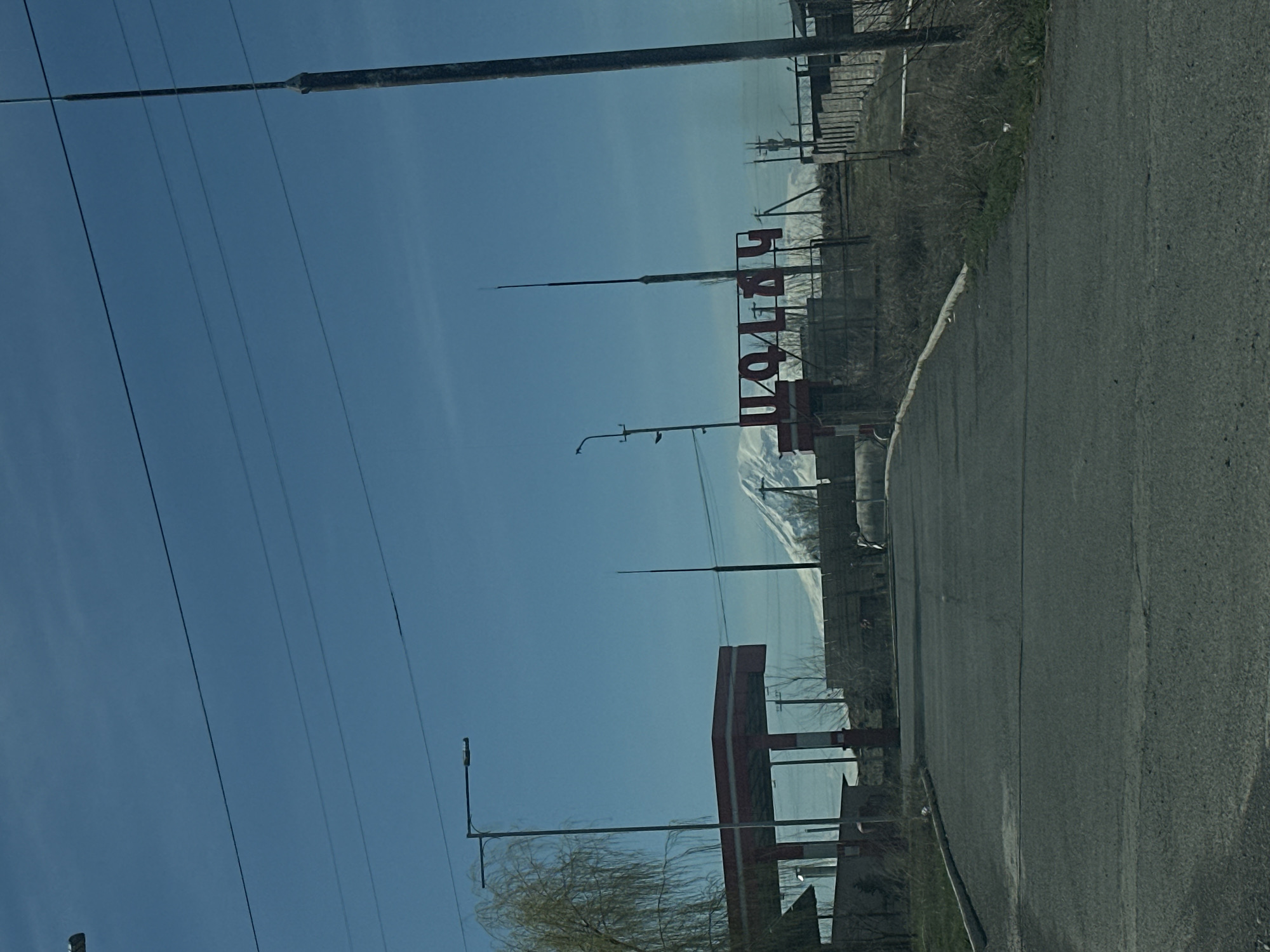 The image shows a road leading into a city. On the right side of the road is a sign in red letters. Behind the sign, snow-capped mountains are visible in the distance. The location of the city is Unknown, Unknown. Overhead, there are power lines strung between poles. A lamppost is visible on the left side of the road. The road itself is asphalt.