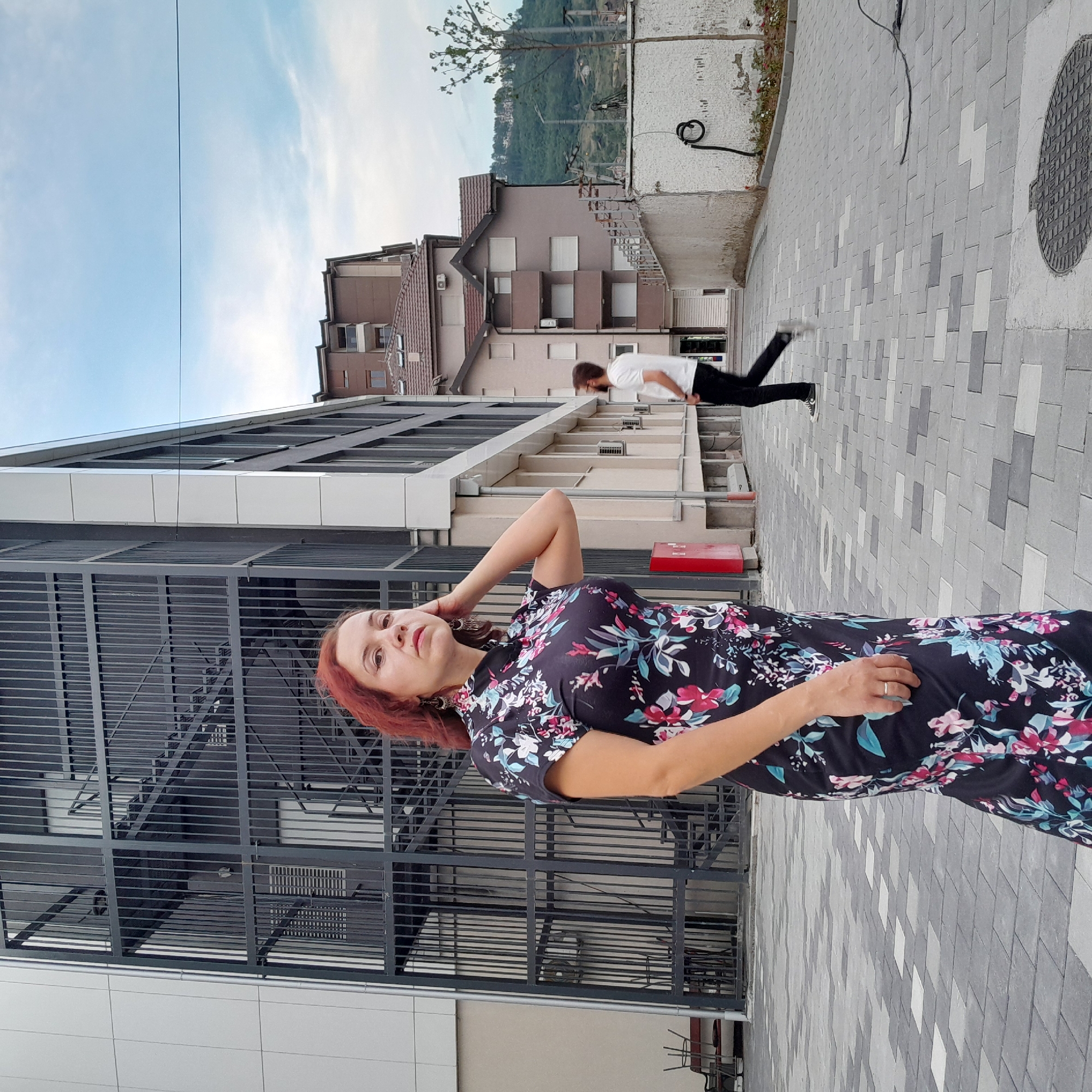 A woman with red hair and a floral dress is lying down and posing for the camera. Her left arm is bent behind her head. She is wearing a ring on her left hand. She has on large, dark earrings. In the background, a man in a white t-shirt and black pants is in mid-air, appearing to jump. He is wearing glasses and dark sneakers. The scene is set outdoors, on a gray, patterned tiled surface. Behind the man, there are stairs leading up to buildings. The buildings appear to be residential or office spaces, with multiple windows. A tree is visible in the distance, with a green, hilly landscape behind it. The sky is blue with some white clouds. The image is vertically oriented.