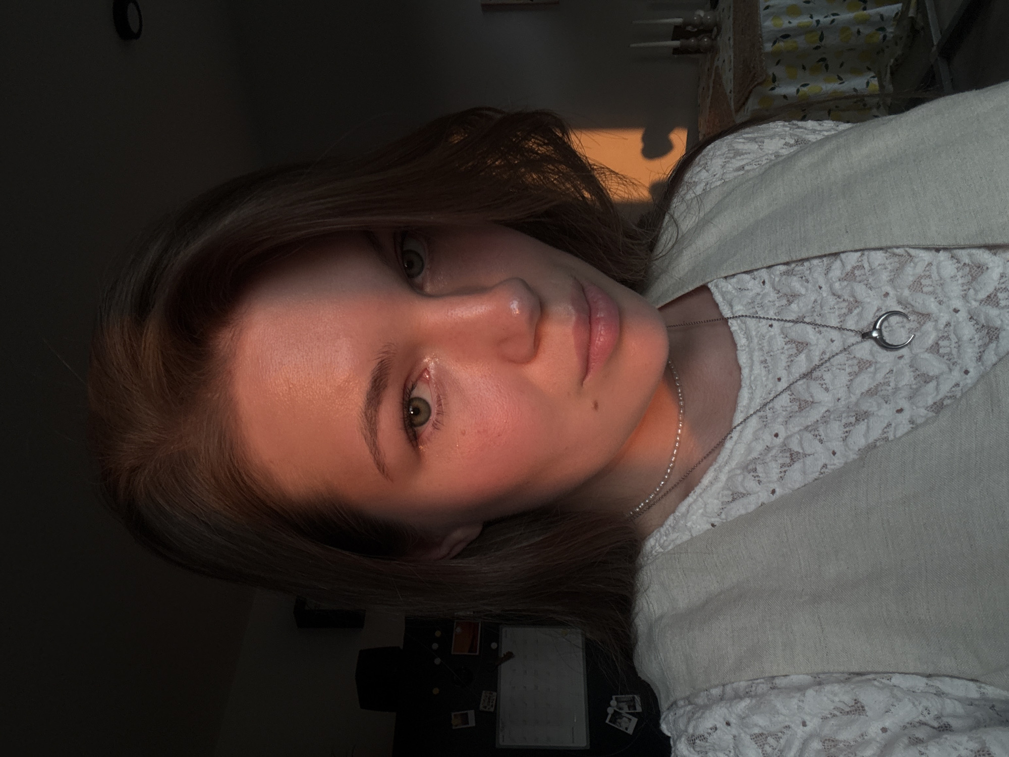 A close-up, slightly angled, indoor portrait of a woman with long brown hair. She is wearing a white lace top with a tan vest over it and a silver necklace with a crescent moon pendant. The right side of her face is illuminated by warm light, while the left side is in shadow. Behind her, a dark surface is visible. In the upper right corner, there are hints of a patterned curtain. The location appears to be indoors, but the specific location in Vozdovac Urban Municipality, Serbia cannot be determined from the image.