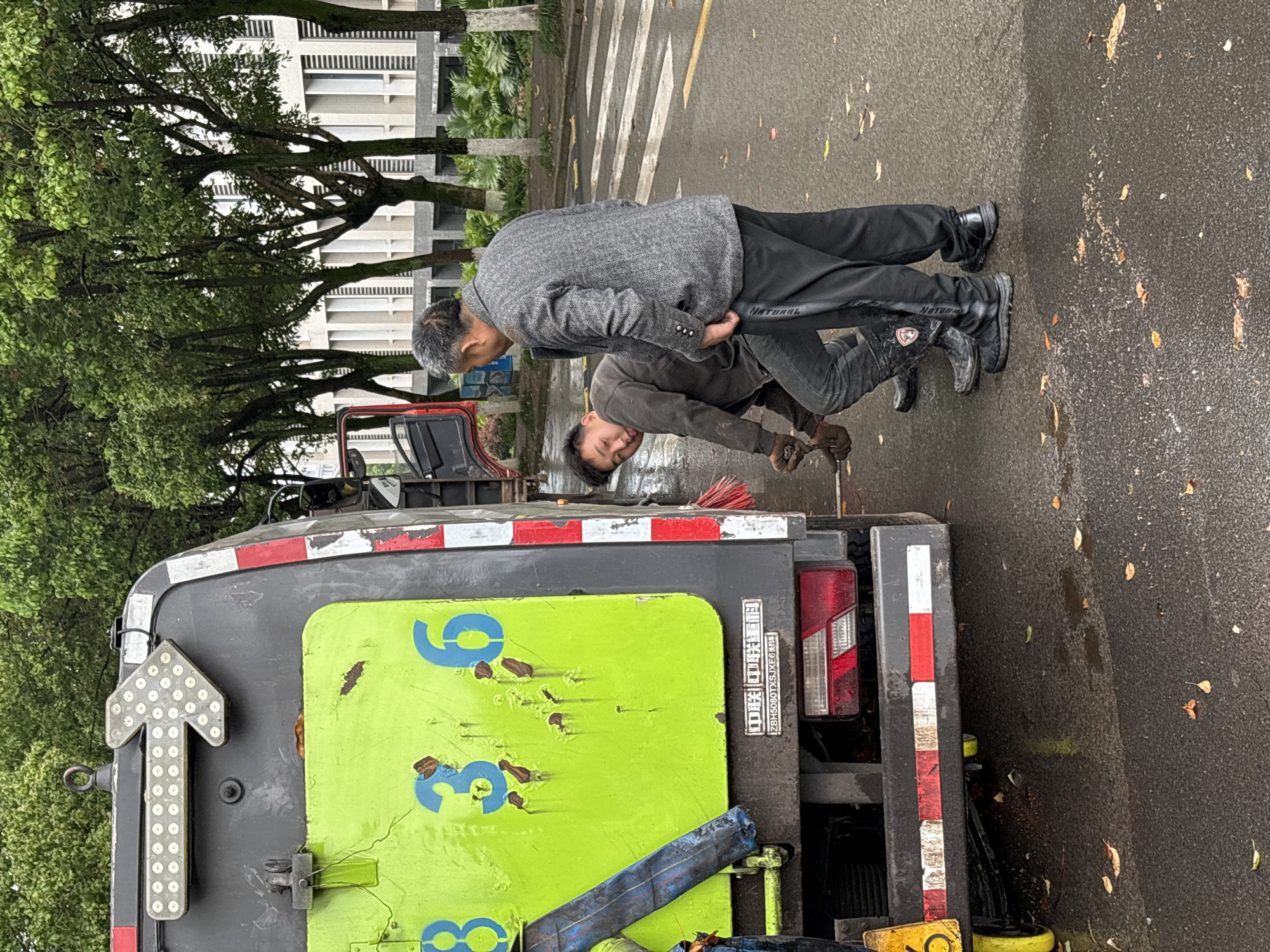 Two men are inspecting the back of a green and gray vehicle with a flashing arrow sign and the number "938" painted on its side. One man is wearing a gray coat and the other is wearing dark clothing and boots. The man in dark clothing is holding a long handled tool, possibly a brush or scraper. The location is Jiangjin, China, as identified by text on the vehicle, and the scene appears to be on a city street, indicated by the painted crosswalk and trees in the background.