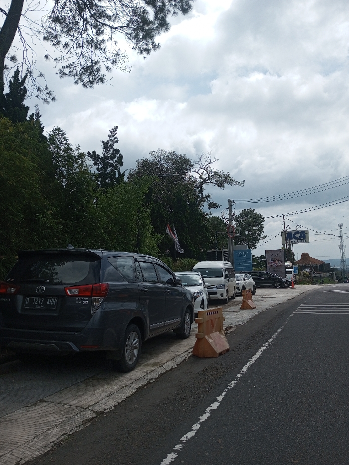 The image depicts a roadside scene in Gubbisamamidi, India, under an overcast sky. A dark grey Toyota Innova SUV, bearing license plate D 1217 AHH, is parked on the far left of the frame, partially on a concrete sidewalk, facing away from the viewer. Behind this vehicle, a white sedan and a white minivan are also parked in a continuous line on the sidewalk. Several orange plastic traffic barriers are strategically placed along the edge of the parking area, delineating it from the asphalt road. The road exhibits a solid white edge line and a partial dashed white lane marker. To the immediate left, a steep green incline densely covered with varied foliage and trees ascends. In the middle and background, multiple utility poles support an array of overhead wires. Prominent commercial signage includes a blue and white "ACA ASURANSI" sign and another darker billboard with white text, partially visible, suggesting "EXPRESSO". A building featuring a distinct peaked roof is discernible in the distance near the "ACA ASURANSI" sign. No individuals are visible within the scene.