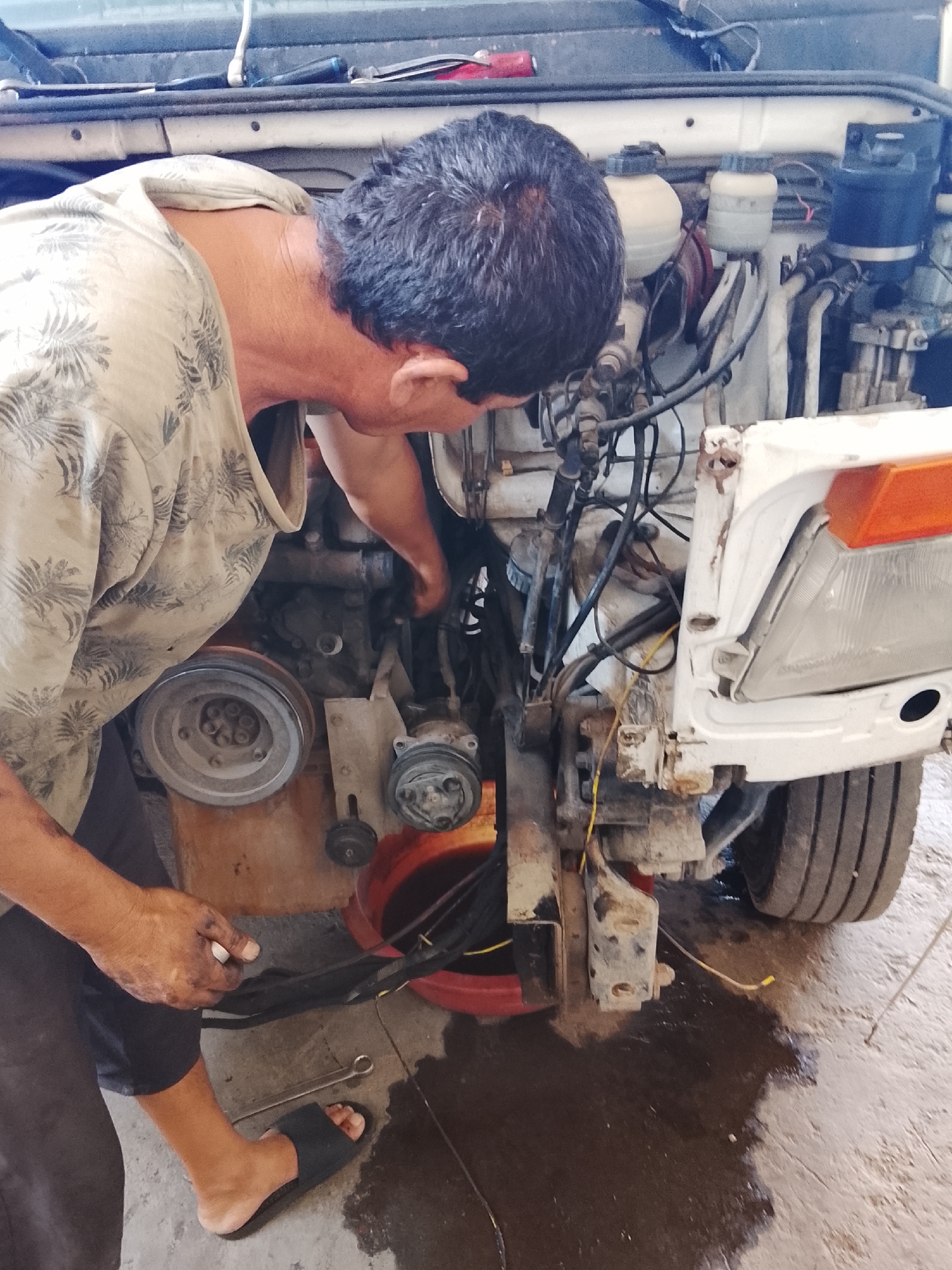 In the city of Sabir Rakhimov, Uzbekistan, a person is seen working on a white vehicle's engine. The individual, wearing a patterned t-shirt and dark pants with a sandal on their left foot, is leaning into the engine bay. Their hands, stained with grease, are actively engaged with the engine's components. A red bucket, likely containing fluids, is positioned near the engine, and a wrench rests on the concrete floor. The vehicle's front end is partially disassembled, revealing the engine, hoses, and wiring. The ground beneath the vehicle is wet, suggesting a fluid leak or recent maintenance. The overall scene depicts a mechanic or vehicle owner performing repairs in an outdoor or garage setting.