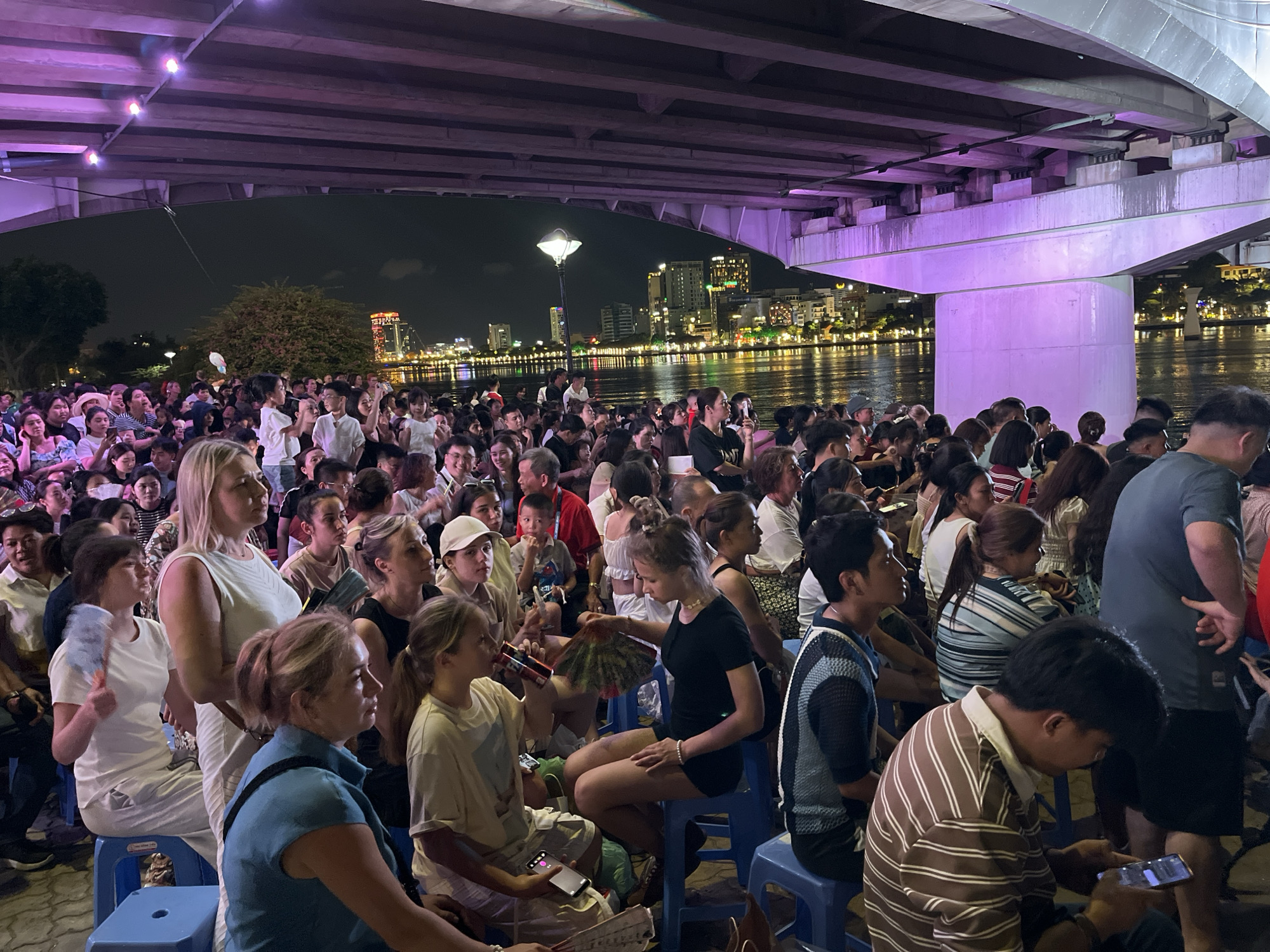 A dense crowd of people, predominantly adults, is gathered in an outdoor setting in Đà Nẵng, Vietnam, likely for an event or performance. The crowd is positioned under a bridge structure, with the city skyline and waterfront visible in the background across the river. Individuals are either sitting on small blue plastic stools or standing, watching something out of frame. Many people are focused on their phones, some using small fans. The bridge overhead is illuminated with purple lighting, creating a dim, low-light ambiance. The general atmosphere suggests an evening event or gathering along the Han River waterfront.