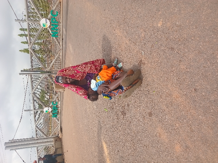 The image shows a woman and a young child sitting on the ground in front of a metal gate. The woman is wearing a red and white headscarf and is holding the child in her arms. The child is wearing an orange shirt and blue pants. The gate is closed, and there is a sign on it that reads "NNPC". The scene is in front of the NNPC Gate in Ungwan Tanko, Nigeria, located at 10.422° N, 7.49° E. The environment is dusty and dry, and the ground is made of gravel. The woman and child appear to be waiting or seeking assistance. There is a man visible in the background, but there is no interaction between him and the woman and child.