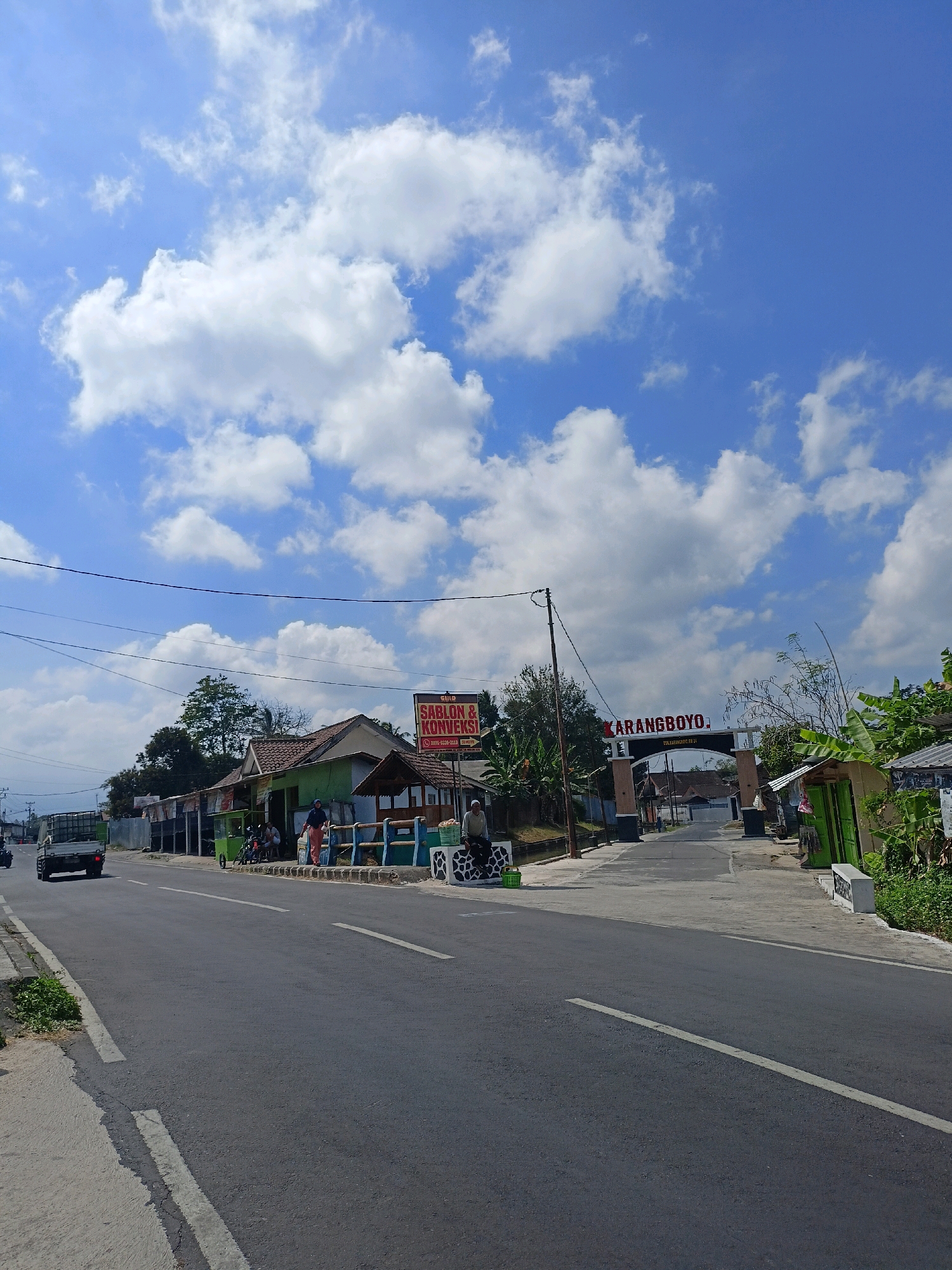 The image shows a street scene in Payaman, Indonesia. On the right side of the road, there is an archway with the text "KARANGBOYO" written on it. To the left of the archway is a building with green trim and a thatched roof. In front of this building, there is a group of people, some standing and some sitting on a low wall. Further down the road on the left, there are several shops with awnings. A truck is driving away from the camera on the left side of the road. The sky is blue with scattered white clouds. The overall environment suggests a rural or semi-urban area with local businesses and residences lining the road.