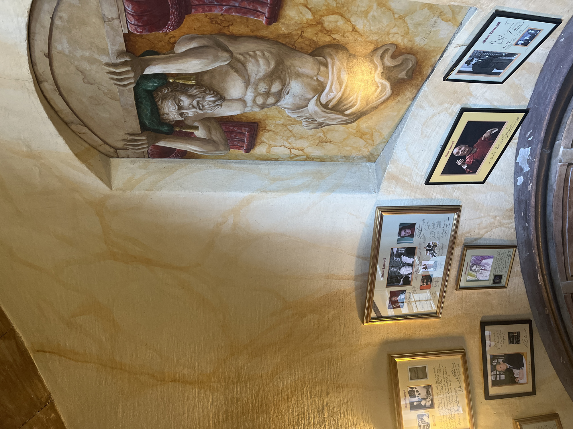 This media file captures an interior scene within a historic building, likely Maison Kammerzell in Strasbourg, France, as indicated by visible text. The focal point is a large, classical-style mural on a vaulted ceiling or upper wall, depicting a muscular, bearded male figure. The figure, possibly a titan or mythological hero, appears to be struggling to hold up a heavy, curved architectural element, rendered in shades of beige and brown with touches of green. The mural's background features a warm, marble-like texture.
On an adjacent curved wall, numerous framed photographs and documents are displayed. These frames, often with gold edges and black mats, hold collections of smaller portraits, both black and white and color, along with handwritten inscriptions and signatures. The text "Maison Kammeryell" (a phonetic spelling of Kammerzell), "Saison Kammeryell," and "Michel Bouguet" are legible, along with a date "1957" and other unreadable dedications. The collection suggests a display of notable patrons, visitors, or figures associated with the establishment.
The walls are painted in warm, textured tones of ochre and yellow. The overall impression is one of an old, richly decorated space with a significant history, illuminated by an unseen light source that creates reflections on the framed glass and highlights the mural's details.