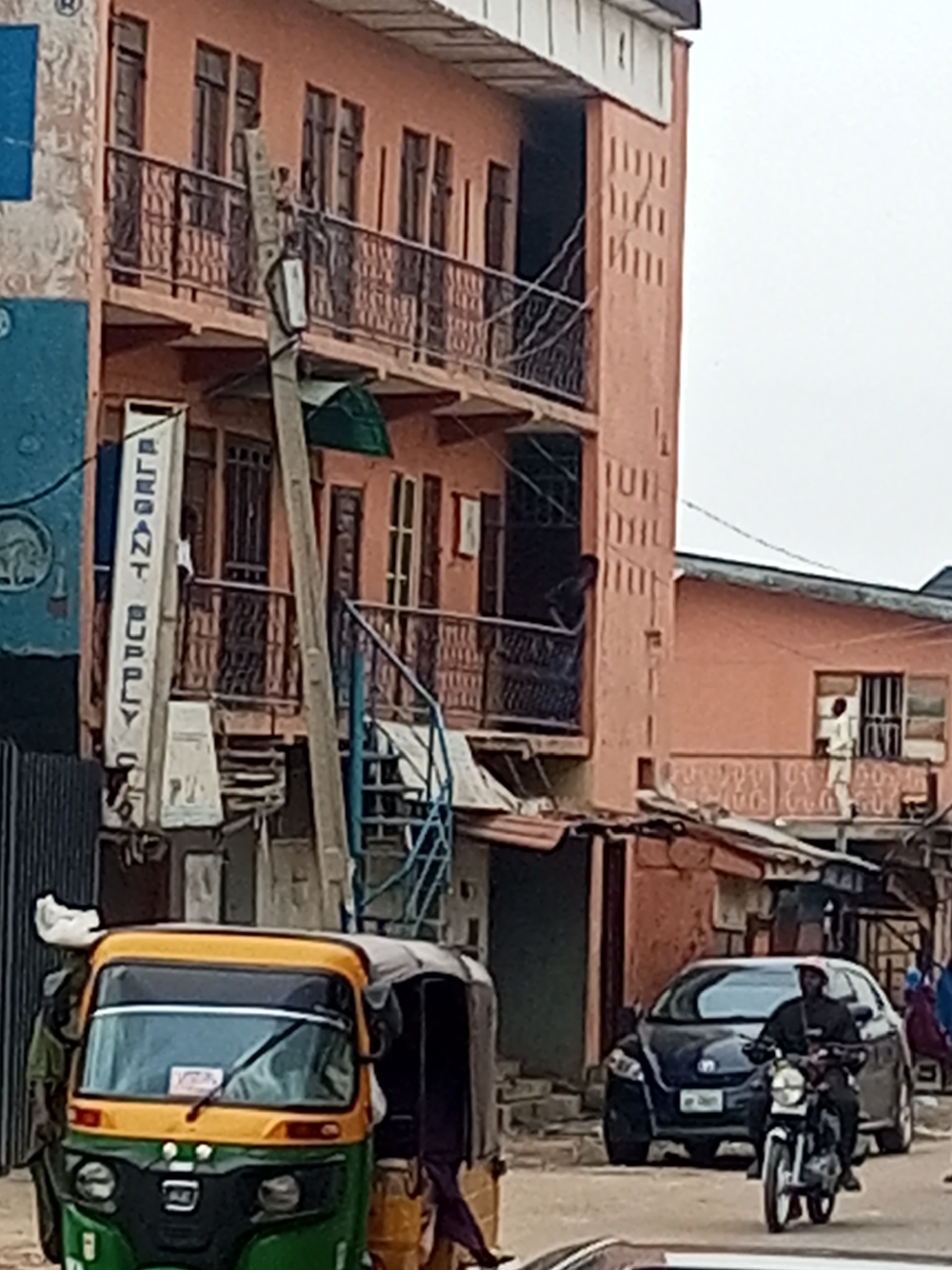 The image depicts a street scene in Maiburiji, Nigeria. In the foreground, a green and yellow three-wheeled vehicle, commonly known as a "Keke Napep," is partially visible. A motorcycle is moving on the road, its rider wearing a black outfit. Behind the motorcycle, a dark-colored car is parked. The background features a multi-story peach-colored building with wrought-iron balconies. A vertical sign with the text "ELEGANT SUPPLY" is attached to the building. Several individuals are visible on the balconies and in the building's windows. Overhead, a network of electrical wires is strung between the buildings. The street appears to be bustling with activity, as indicated by the presence of vehicles and pedestrians.