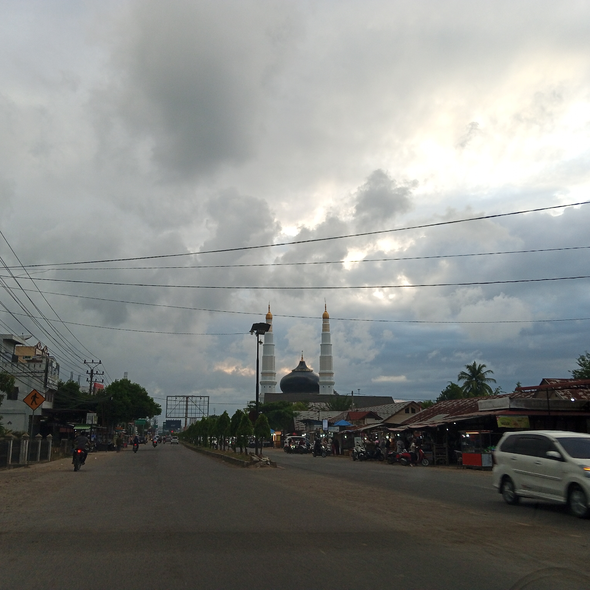 This image captures a street scene in Pantegajah, Indonesia, under an overcast sky with patches of brighter light visible through the clouds, indicating either sunrise, sunset, or partial clearing. The central background features a prominent mosque with a large, dark, possibly black or dark green dome, flanked by two tall, white minarets topped with gold-colored conical spires.
In the middle ground, an asphalt road stretches into the distance. Several vehicles are present: a white multi-purpose vehicle (MPV) or minivan is observed on the right side of the road, partially blurred due to motion, suggesting it is moving towards the viewer. Further down the road, multiple motorcycles are visible, with at least one rider in the foreground on the left, moving away. On the left side of the road, there are buildings, power poles, and a dense network of overhead electrical and communication wires spanning across the upper half of the image. A yellow pedestrian crossing sign is visible on the left.
The right side of the road features a row of small, tree-like plants lining the median, followed by various roadside structures that appear to be small shops or stalls, some with corrugated metal roofs and illuminated interiors, suggesting commercial activity. People are gathered around these stalls, some on motorcycles, indicating typical street market or daily commuter activity. Palm trees and other vegetation are scattered throughout the background and along the sides of the road. A street light fixture is visible near the mosque. The overall lighting suggests either early morning or late afternoon, given the atmospheric conditions.