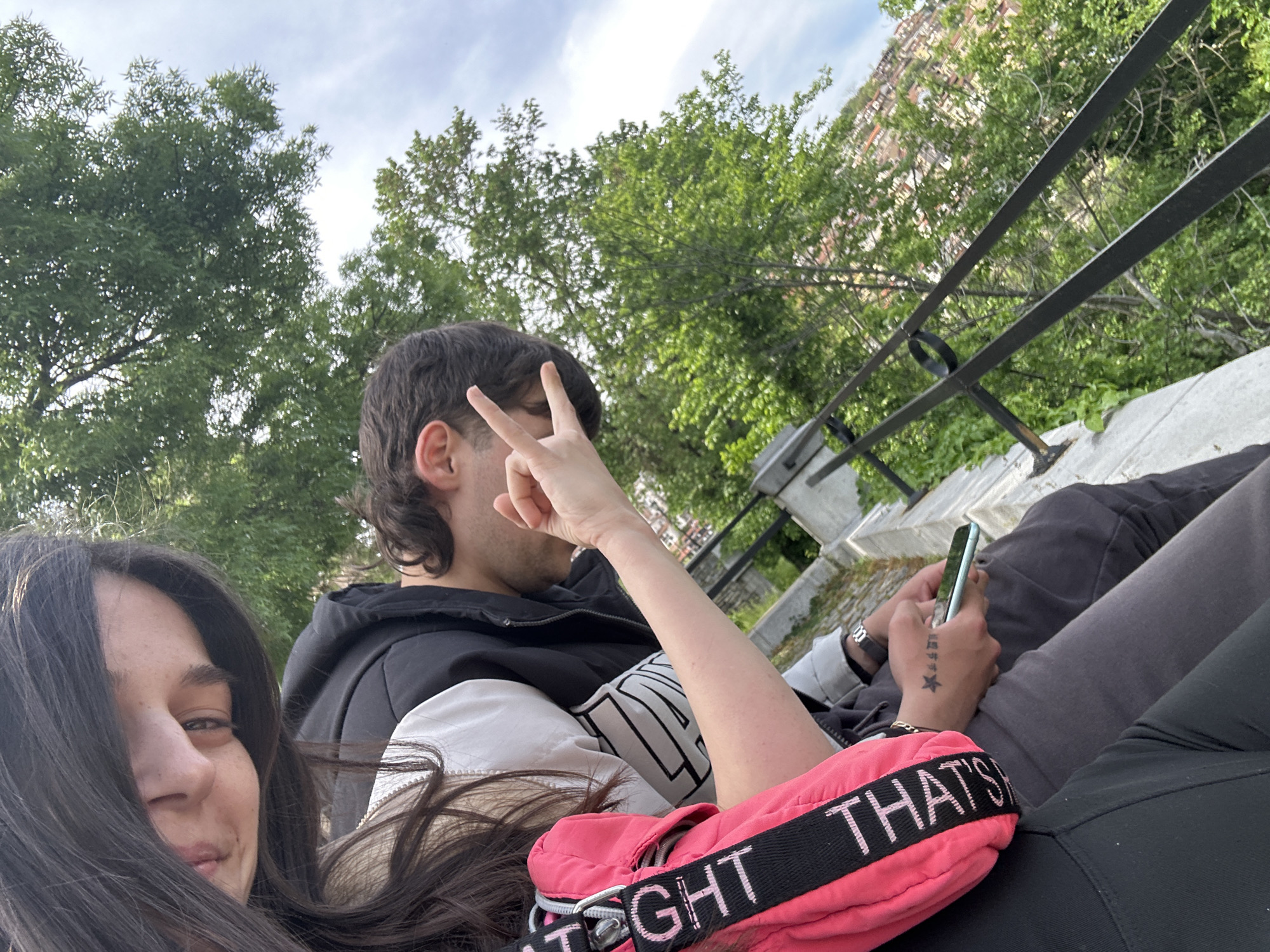 The photograph, taken at an upward angle, depicts two individuals in an outdoor setting. A woman with long, dark hair is visible in the lower left corner of the frame, her face turned toward the camera. To her right, a man is seated, making a peace sign with his left hand. He has dark hair and is wearing a black and white jacket. He is holding a cell phone.
A pink fanny pack is present. The pair are seated on a low wall or ledge. A black metal railing is visible. In the background are green trees and a building that could be in Veliko Tarnovo, Bulgaria. The sky is visible at the top of the frame, with scattered clouds.