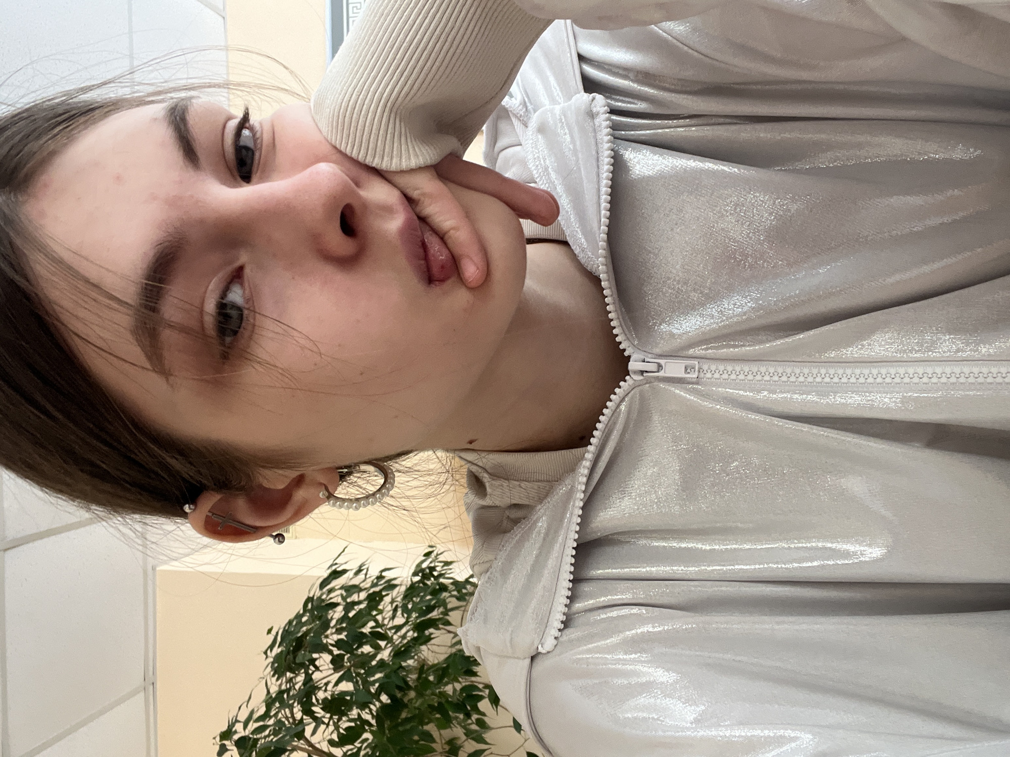 A close-up, tilted, eye-level shot shows a young woman. She is fair-skinned with dark hair and eyes. She is wearing a metallic silver jacket with a white zipper and a white ribbed sweater underneath. Her left hand is resting on her cheek, and her tongue is slightly visible. She is wearing hoop earrings and multiple piercings on her left ear. In the background is a green plant, a light-colored wall, and a partially visible window frame. There is no information to verify that this photo was taken in Saint Petersburg, Russia.