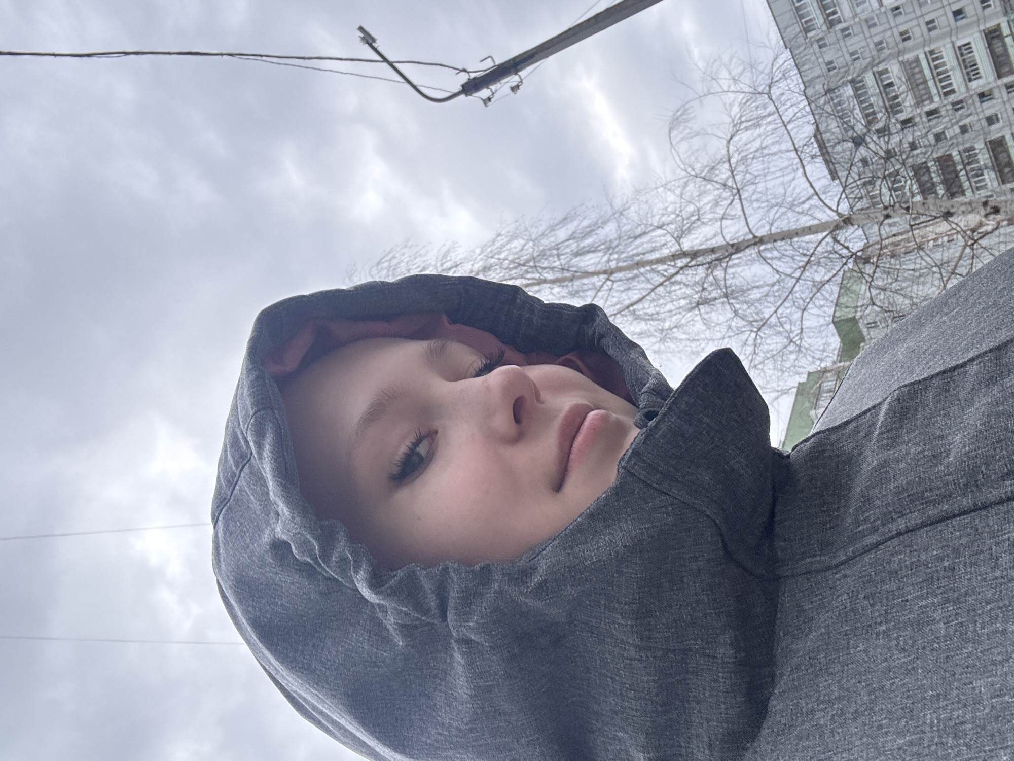 A low-angle close-up shows a person wearing a gray hooded jacket. The hood frames their face, and the lining of the hood appears to be a dark red or maroon color. The person's skin tone is fair, and only one eye is fully visible. The sky is overcast and cloudy. In the background, there is a utility pole with wires, bare tree branches, and a multi-story building. The location is Chaikovsky, Russia.
