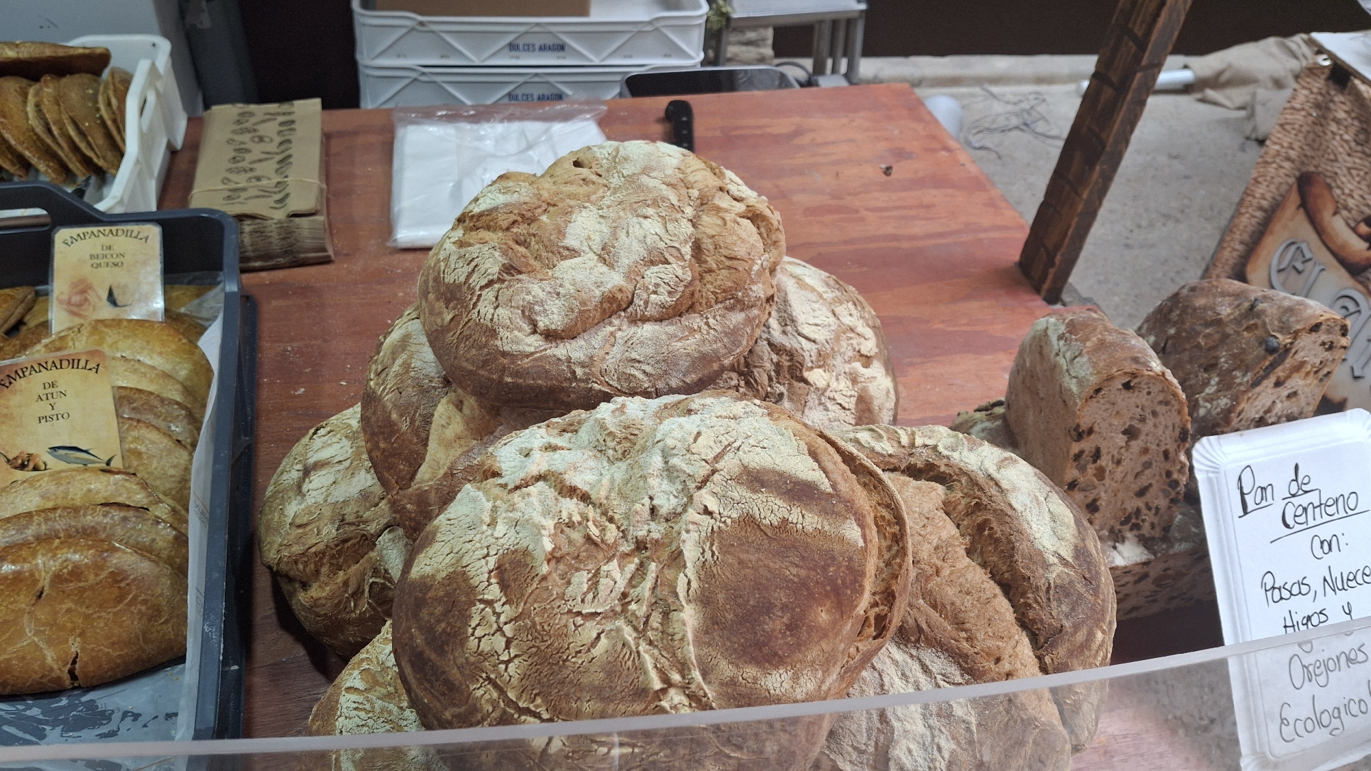 The image depicts a market stall in La Fresneda, Spain, displaying a variety of freshly baked goods. In the foreground, several round loaves of bread with a rustic, crusty exterior are piled up. To the left, a black plastic tray holds individual empanadas, some with descriptive labels. One label reads "Empanadilla de Bercon y Queso" and another "Empanadilla de Atun y Pisto." Further back, another white tray contains more empanadas.

To the right of the bread, a loaf of dark rye bread, identified by a handwritten sign as "Pan de Centeno Con: Pasas, Nuez, Higos y Orejones Ecologico!", is visible. This rye bread appears to contain dried fruits and nuts. Behind this loaf, another dark bread with visible inclusions is present.

The stall itself is set up on a wooden surface, with a rustic wooden beam structure supporting a backdrop. A woven basket is visible to the far right. The overall impression is one of a local food market offering artisanal baked products. No people are visible in the image.