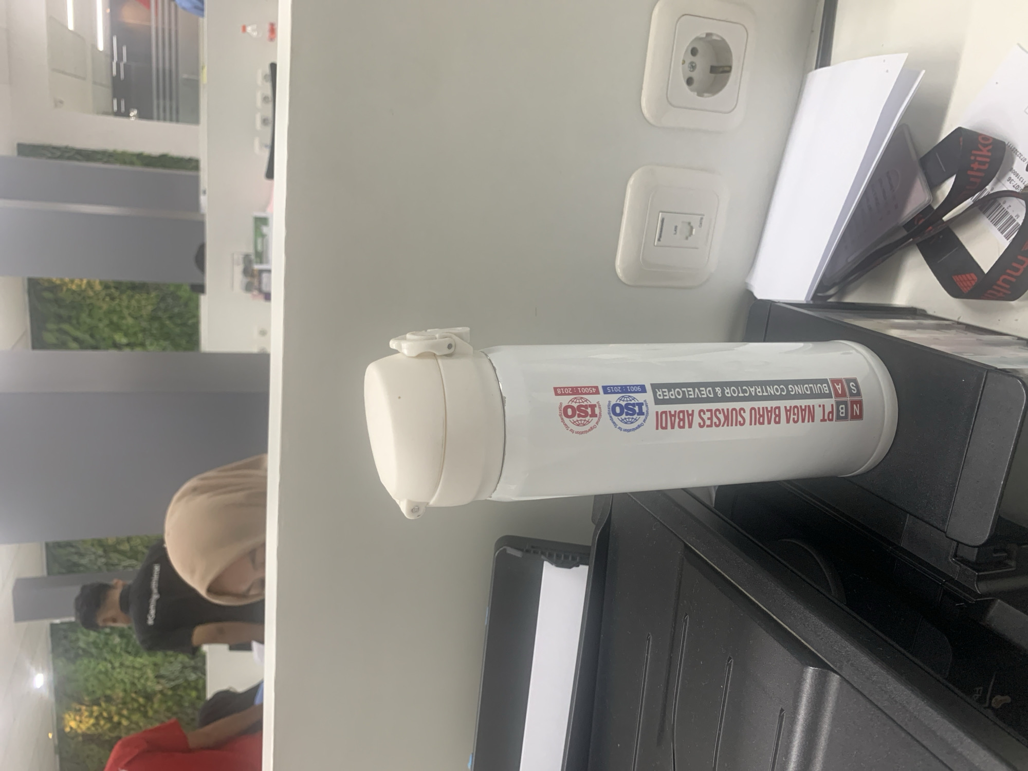 The image depicts an indoor office environment. In the foreground, a white thermos with blue and red text is positioned on a black printer. The text on the thermos reads "N B PT. NAGA BARU SUKSES ABADI S A BUILDING CONTRACTOR & DEVELOPER" along with ISO certifications. To the left of the thermos is a black printer. In the background, a person wearing a light brown hijab is partially visible. Further in the background, another person in a black t-shirt is seen from the back, and a person in a red shirt is on the far left. The walls feature green vertical plant installations and grey panels. Electrical outlets are visible on the wall behind the thermos. The image does not provide any specific information about the city of Palembang, Indonesia.