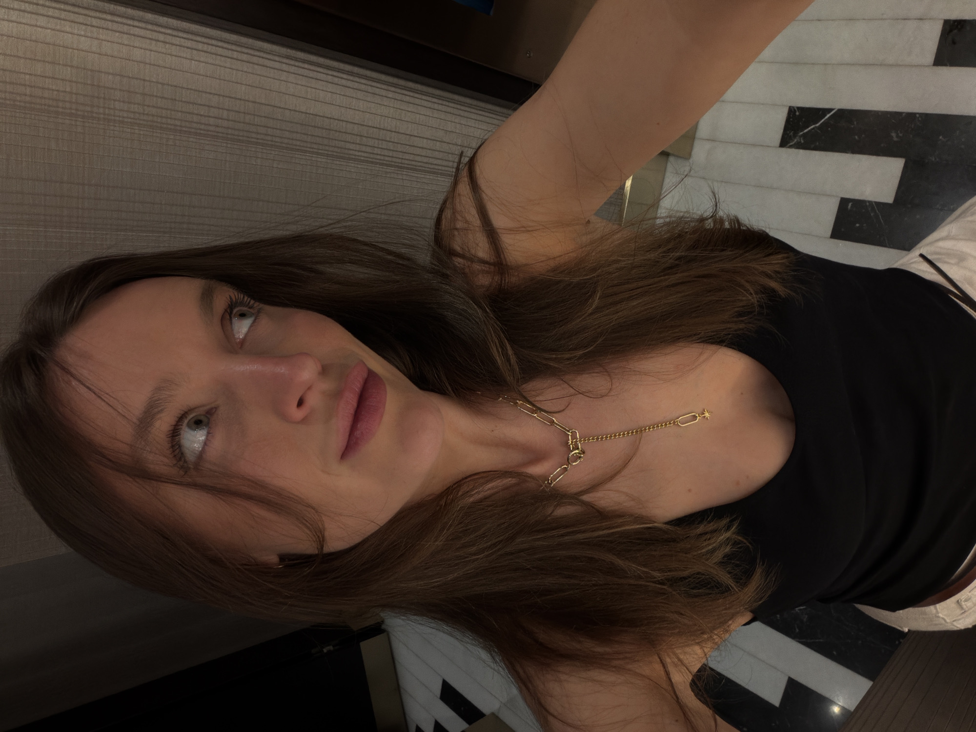 The image is a selfie of a woman indoors, likely in a hotel or apartment. The background features a wall with horizontal stripes and a floor with alternating black and white rectangular tiles. The woman has long brown hair and is wearing a black top and a gold necklace. She is looking up and to the left of the frame. The location is possibly Budva, Montenegro, although there is no conclusive evidence in the image to confirm this. There are no other people or subjects visible. The primary action is the woman taking a selfie.