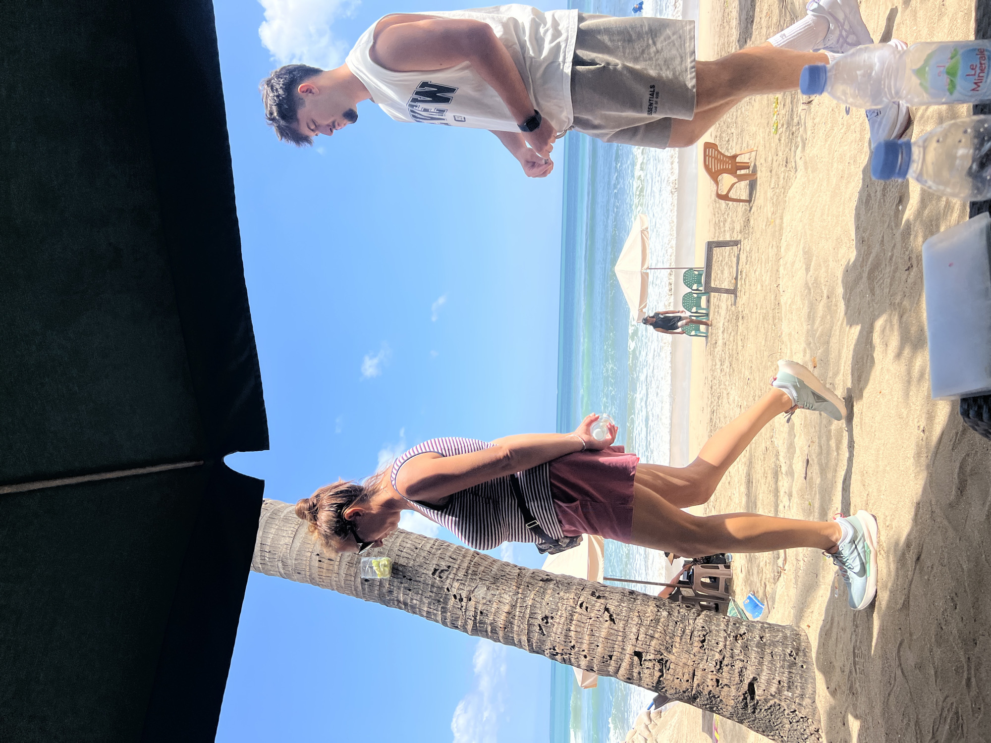 The image depicts a sunny beach scene in Kuta, Indonesia, with the ocean and blue sky dominating the background. In the foreground, a woman with her hair in a bun, wearing sunglasses, a striped tank top, and pink shorts, walks from left to right, holding a clear plastic cup with a green beverage and a bottle of water. To her right, a young man with dark hair and a goatee, wearing a sleeveless off-white t-shirt and grey shorts, stands looking to his left. A large palm tree trunk is visible on the left side of the image. In the mid-ground, beach chairs and umbrellas are set up on the sand, and two people are visible near the water. The sand is light brown, and the ocean waves are breaking gently. The scene is under the shade of a large dark umbrella.