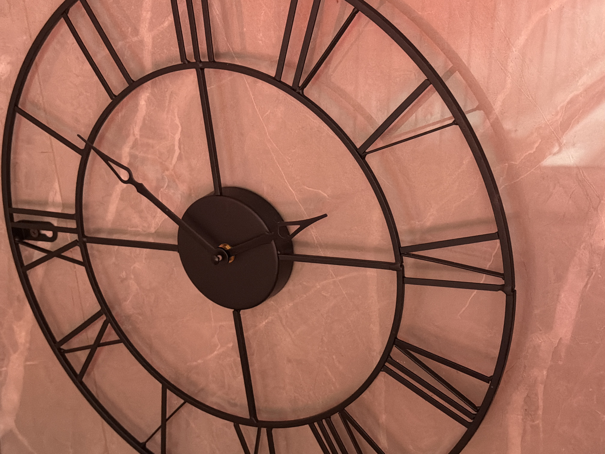 This media file features a close-up, angled view of a large, black, wrought-iron wall clock with Roman numerals. The clock face is set against a textured, rose-colored background, possibly a wall with a marble or stone finish. The clock has two hands: a shorter hour hand and a longer minute hand. The hour hand points slightly past the Roman numeral IX, and the minute hand points directly at the Roman numeral VI. This indicates the time is approximately 9:30. There is a subtle shadow cast by the clock on the wall behind it, suggesting a light source from the side. The overall impression is one of an interior setting, possibly a home or a decorative space, with a focus on the clock as a decorative object. No people are visible in the frame. The lighting is warm, creating a soft ambiance. The image does not contain any visible text. The location context provided is Orenburg, Russia, but there are no specific visual cues in the image to confirm this location.