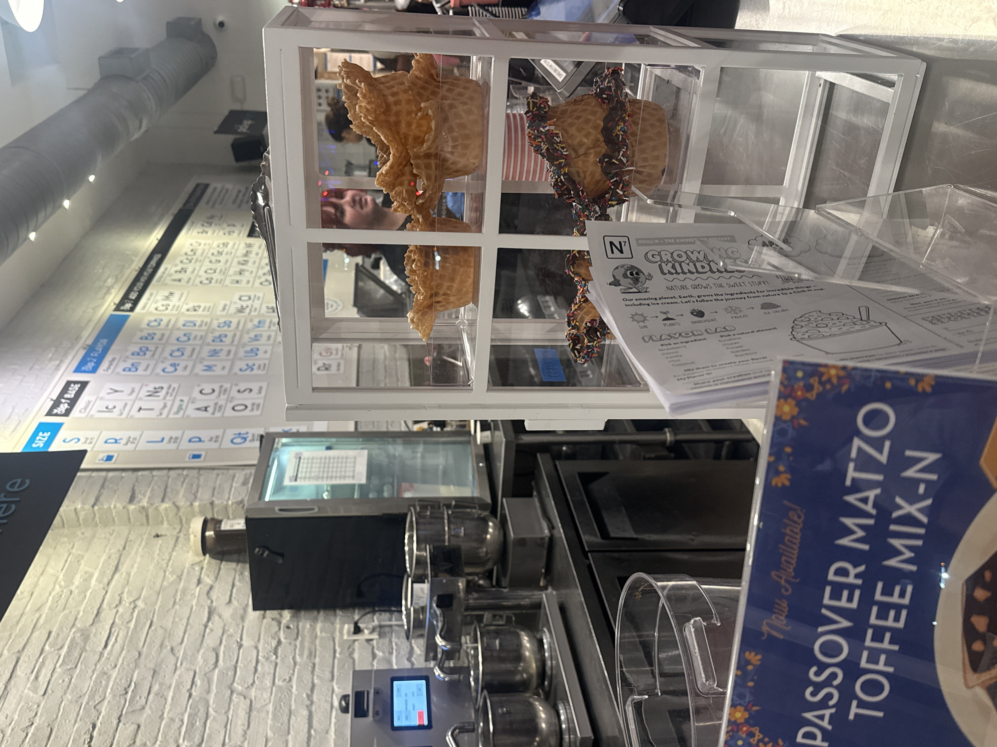 The image shows the interior of a dessert shop in Aventura, Florida, United States. A white metal display case contains decorated waffle cones. A customer is visible behind the case. In front of the display are promotional materials for "Growing Kindness" and "Passover Matzo Toffee Mix-N." A periodic table of flavorings hangs on the wall. Professional ice cream preparation equipment is next to the counter.