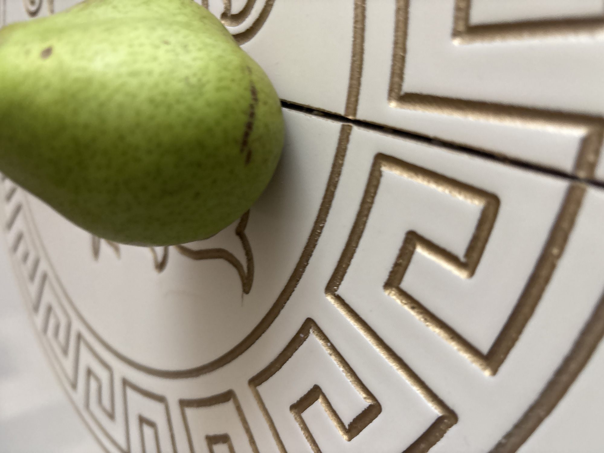 The image is a close-up shot of a green pear resting on a white surface with a gold geometric pattern. The pattern appears to be a repeating Greek key design. The lighting is bright, casting subtle shadows. The focus is on the pear and the textured surface, with the background slightly blurred. There is no discernible location in Georgiyevsk, Russia, or any people present in the image.