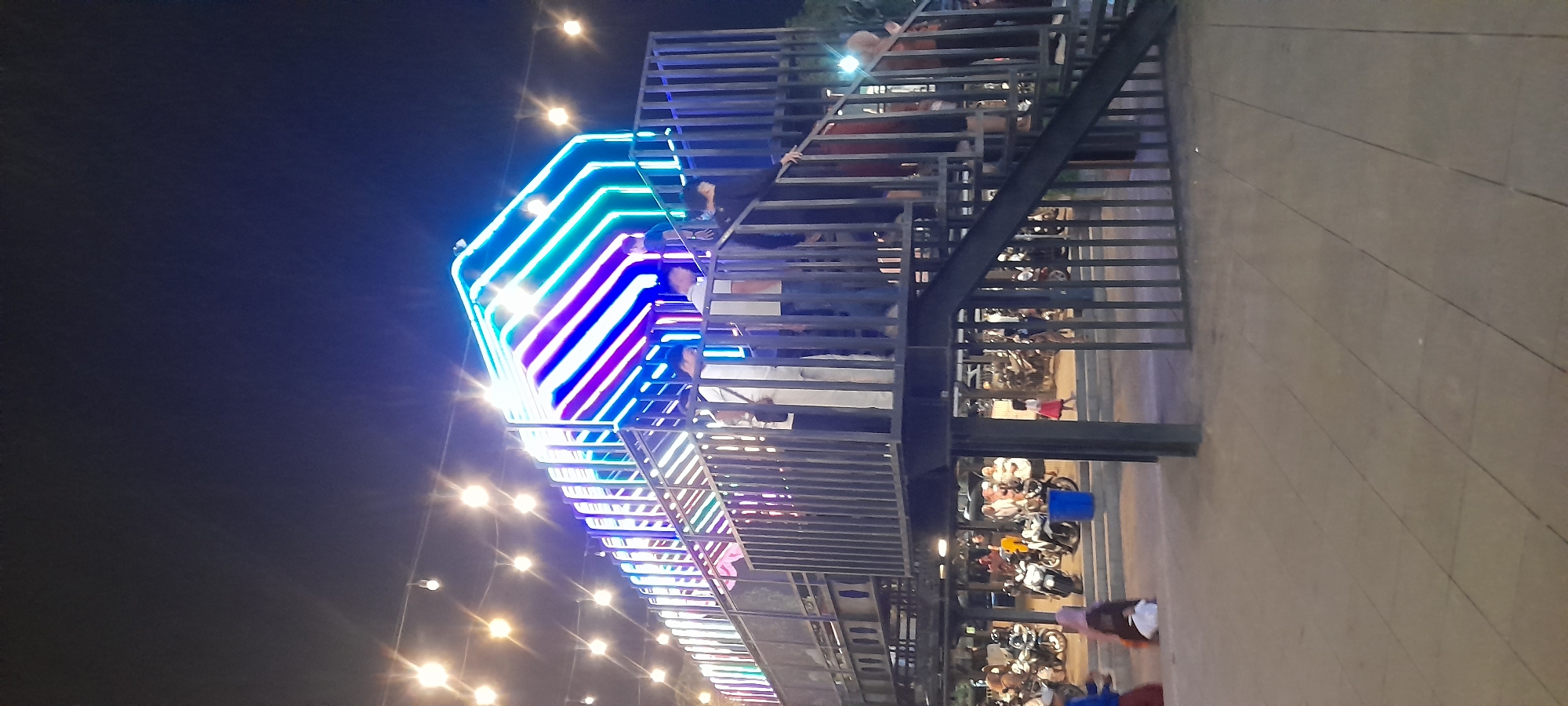 The image shows an outdoor scene at night, likely in a public space in Situbondo, Indonesia, characterized by neon lights and a pedestrian walkway. In the foreground, a dark grey metal staircase with vertical bars leads up to a walkway. Several individuals are visible on the staircase, some appearing to be descending, while others are at the top. The walkway is bordered by similar metal railings. Above the walkway, a structure is illuminated by vibrant neon lights in shades of blue, green, and purple, creating a striking visual effect against the dark night sky. String lights, resembling small bulbs, are also suspended across the scene, adding to the festive atmosphere. In the background, a gathering of people and numerous motorcycles are present, suggesting a public event or marketplace. The ground surface appears to be paved with square tiles.