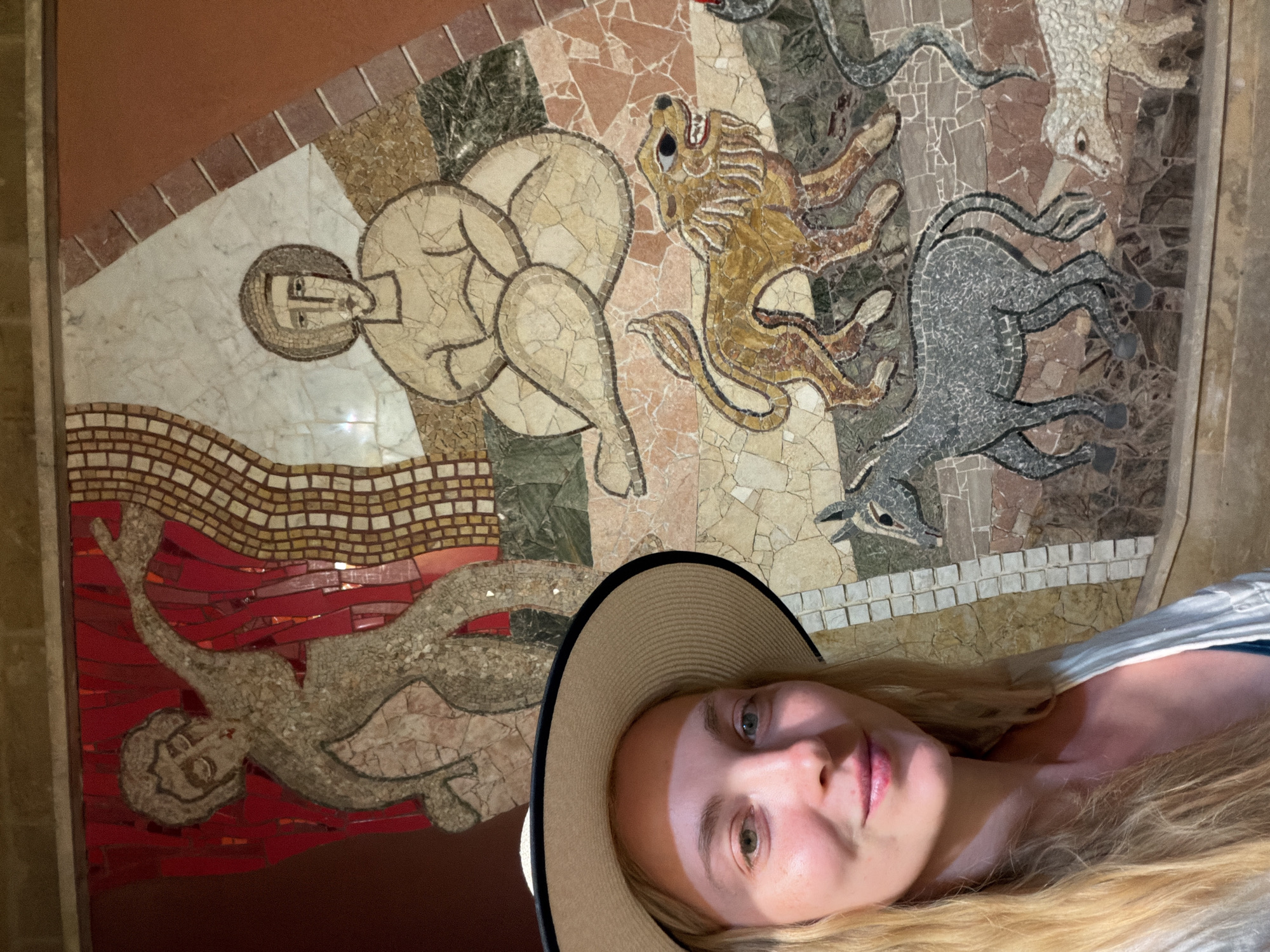 A woman wearing a wide-brimmed hat poses for a selfie in front of a large, colorful mosaic mural in Valletta, Malta. The mosaic depicts stylized human figures and animals, including a lion and a donkey. The woman is smiling and looking at the camera. The mural is composed of small, irregular pieces of tile, creating a textured surface. The background is a warm, earthy tone. The mural is located on a wall in a public space.