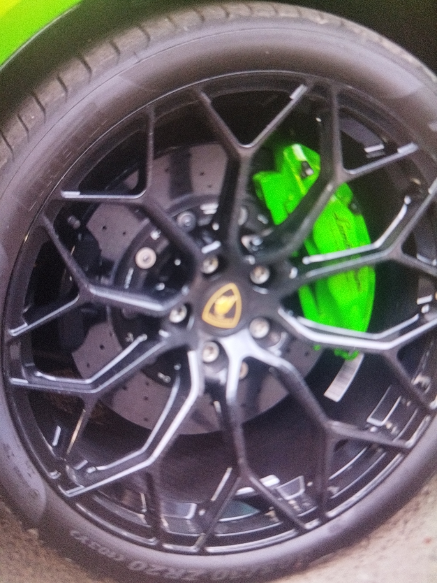 The image shows the black alloy wheel of a Lamborghini sports car. A Lamborghini emblem is visible at the center of the wheel. Behind the wheel spokes, bright green brake calipers are visible, with the "Lamborghini" logo. The tire size appears to be "30 ZR20 (103Y)." The image does not contain enough details to determine the specific location in Plovdiv, Bulgaria. It is a close-up view of the wheel. There are no people visible in the image.