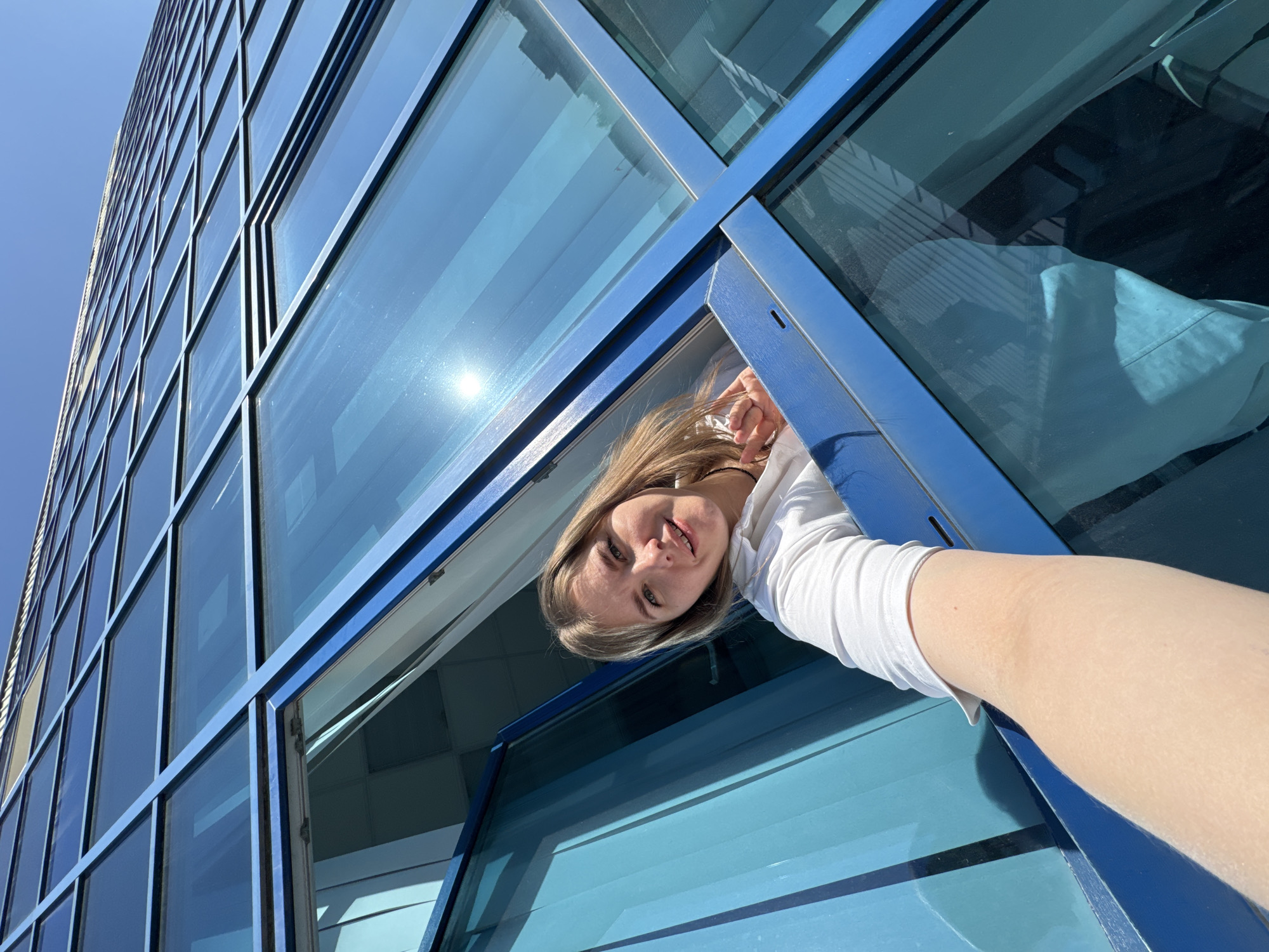 A woman is leaning out of an open window of a tall, modern building in Perm, Russia. The building has a blue-tinted glass facade with a grid-like pattern of metal frames. The window is also framed in blue. The woman has light brown hair and is wearing a white shirt. She is looking towards the camera and smiling. The sky is clear and blue. The sun is reflecting off the glass windows of the building. The angle of the shot is looking upwards from below, giving a sense of the building's height.