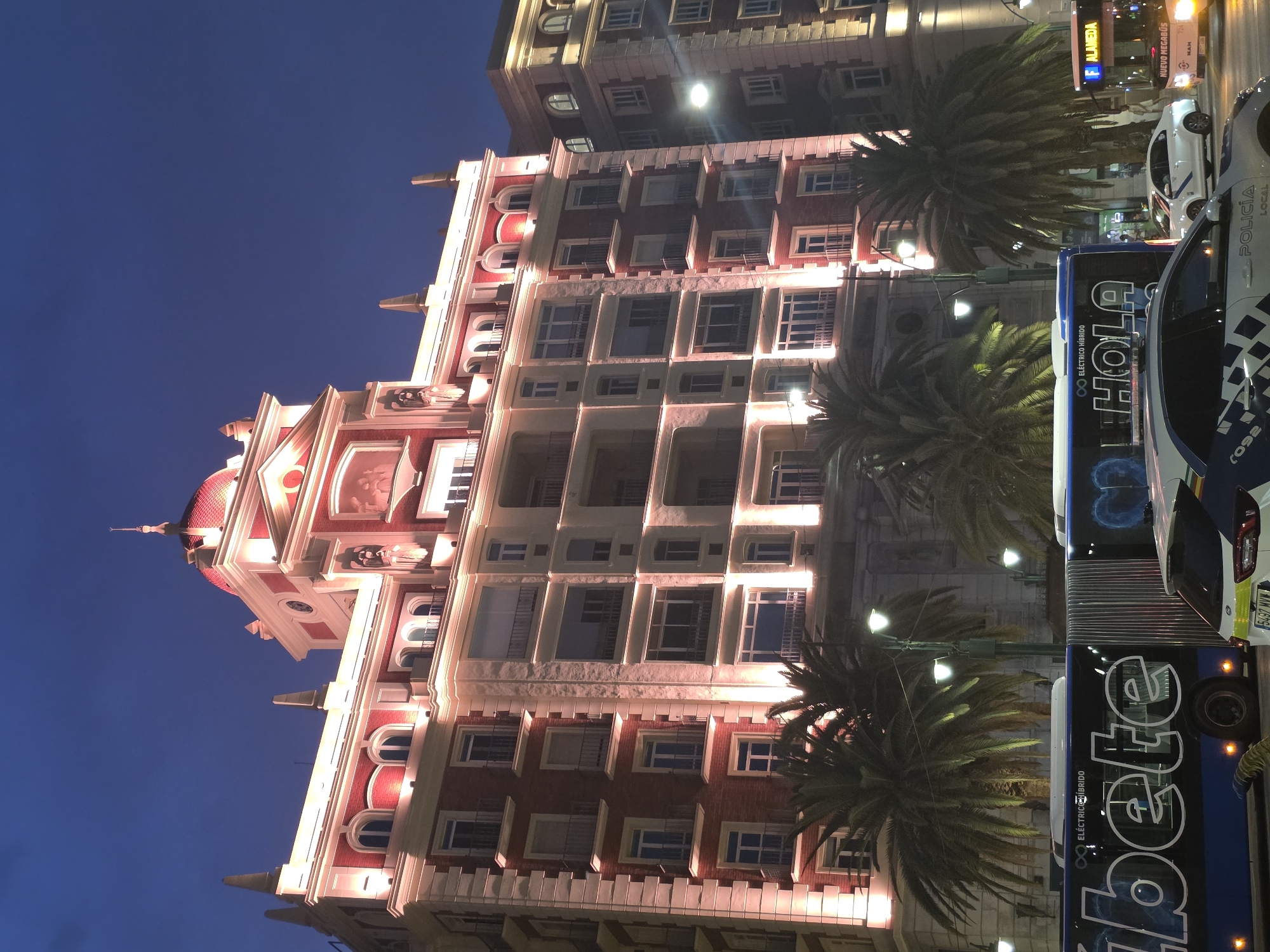 This is a nighttime, low-angle, exterior, full-shot photograph of a grand, multi-story building illuminated by warm, artificial lights. The building features red brick and cream-colored stone architectural elements, including ornate window frames, balconies with black railings, and a prominent dome-shaped roof topped with a spire. The building is situated in Málaga, Spain, likely on the Alameda Principal, as indicated by the bus signage. In the foreground, palm trees with their fronds illuminated by the building lights frame the scene. A blue articulated bus, displaying the word "betete" in large white lettering and "ELÉCTRICO HÍBRIDO" above it, is parked on the left. To the right of the bus, another bus with "F ALAMEDA" and "NUEVO MEGABÚS" displayed is visible. A police car with "POLICIA LOCAL" marked on its side is positioned in the lower right quadrant of the image, with its headlights on and reflecting off the wet street. The sky is a deep twilight blue, suggesting the photograph was taken shortly after sunset. The overall impression is one of urban elegance and nighttime activity.