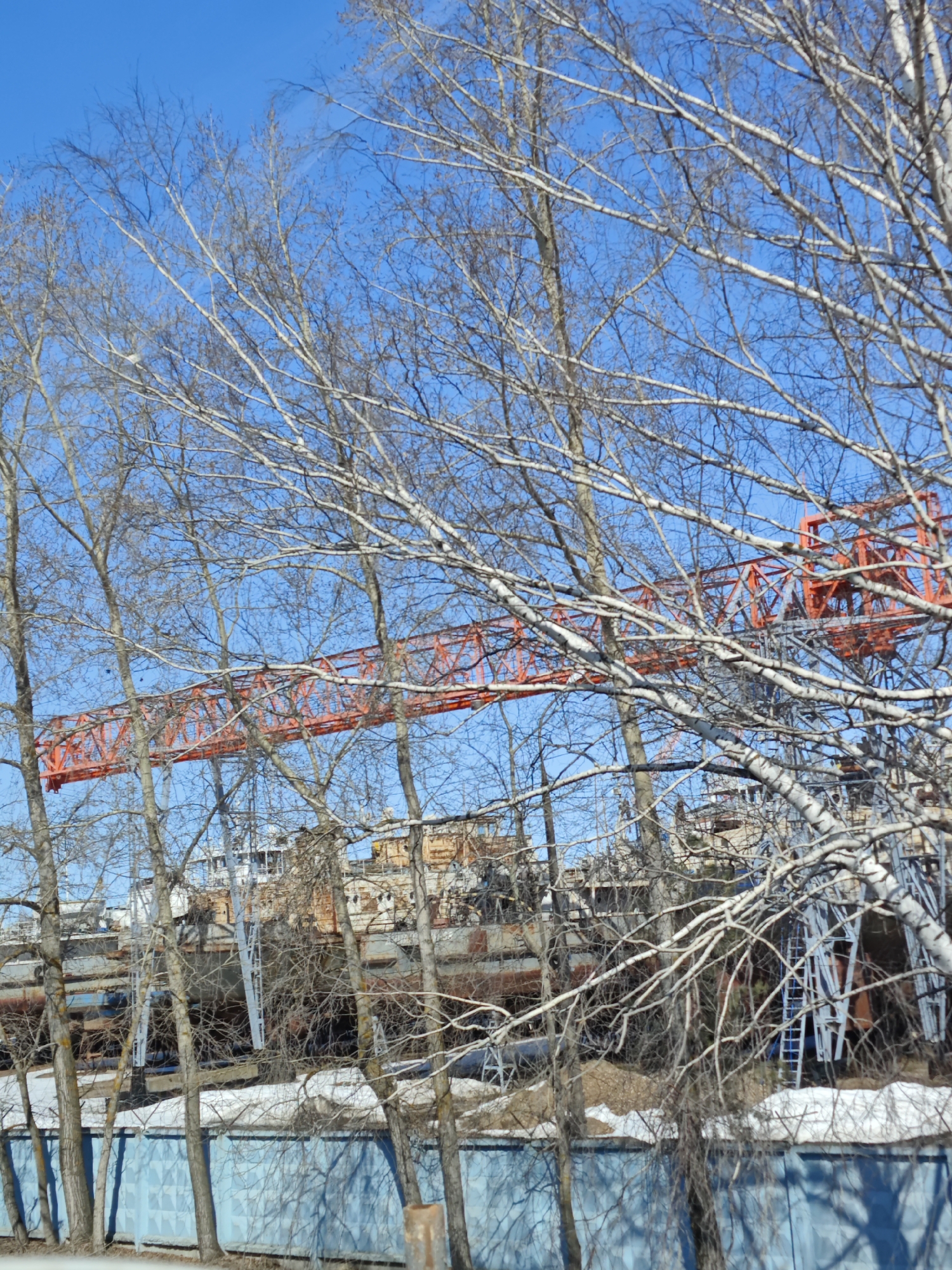 The image shows a view of the Chaikovsky Shipyard in Chaikovsky, Perm Krai, Russia. Several docked ships are visible behind a row of bare trees. An orange overhead crane is also seen through the trees. The foreground shows a light blue fence casting shadows. The sky is clear and blue.