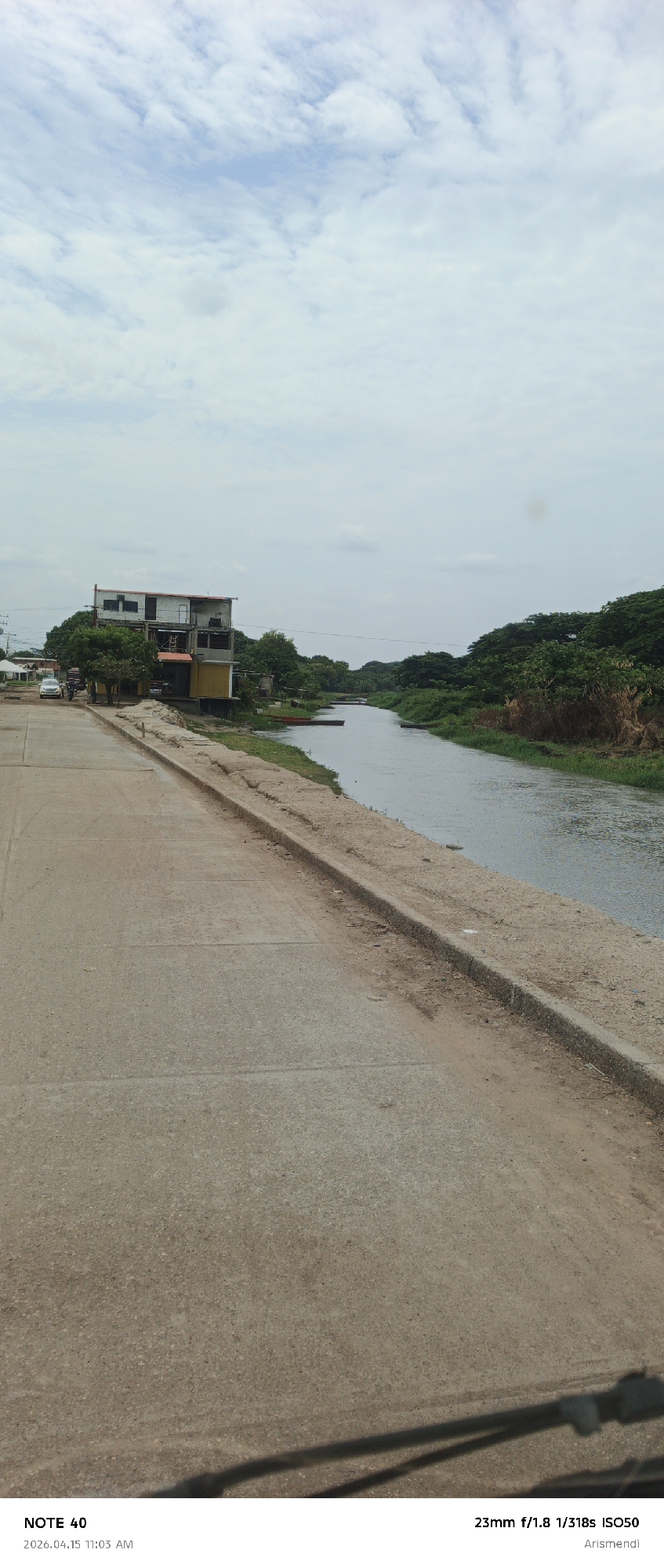 The image presents a view from what appears to be a vehicle, showing a river scene in Arismendi, Venezuela. The river flows from left to right in the middle ground, bordered by vegetation on the right bank and a concrete embankment and road on the left. The left side of the image is dominated by a paved road, and a building can be seen in the background on the left side of the river. The sky is overcast, with a mix of white and grey clouds. The overall scene suggests a rural or suburban setting within Arismendi, Venezuela, near a waterway. There are no visible people or specific actions taking place, other than the potential movement of the vehicle taking the picture.