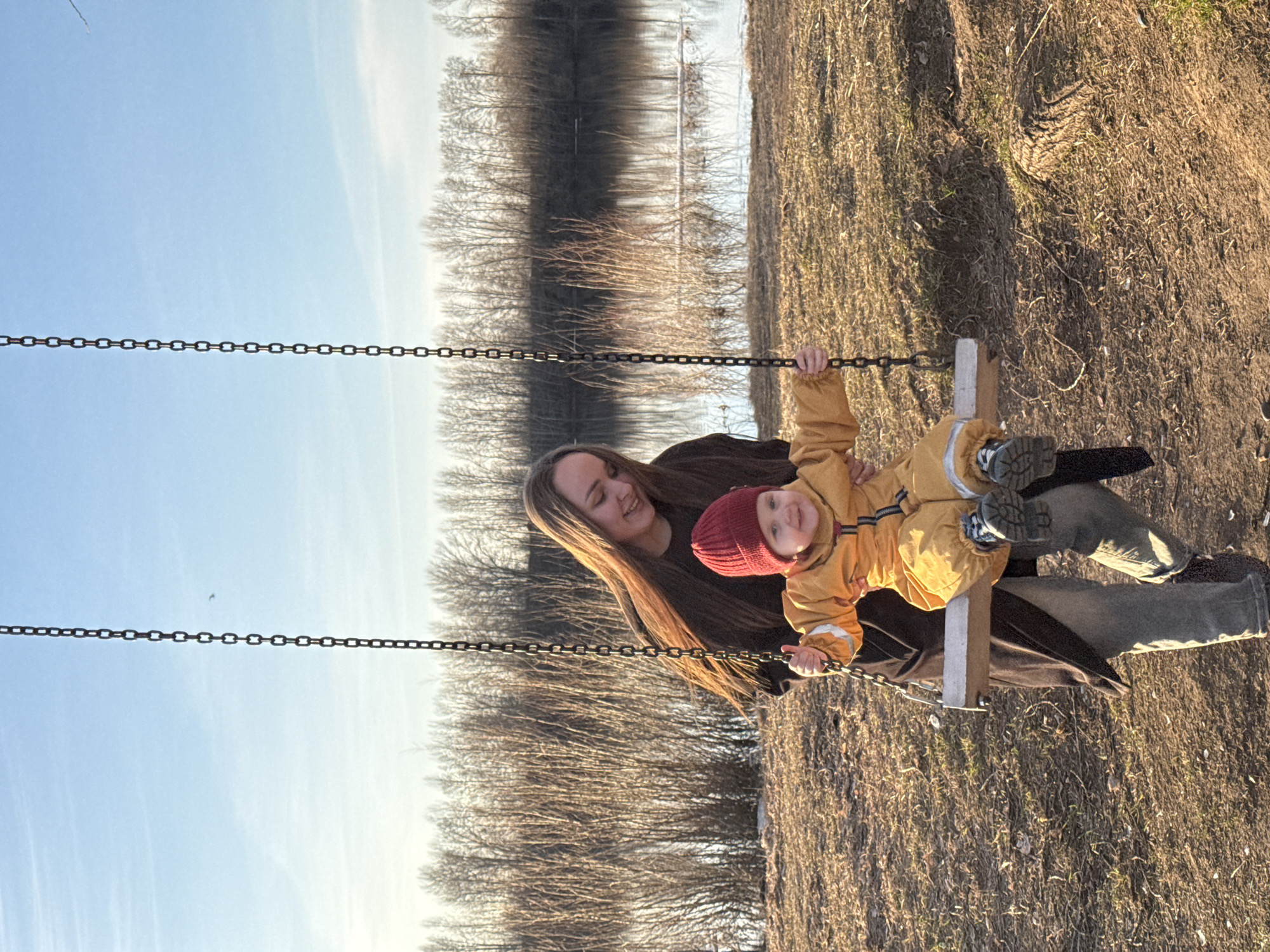 An outdoor scene in Chaikovsky, Russia, depicts a young woman and a child enjoying a swing on a sunny day. The image is composed with the subjects positioned towards the lower right, and the swing chains diagonally across the frame.
A young woman, likely the mother, with long, light brown hair, is seen smiling warmly as she interacts with the child. She wears a dark top or jacket. The child, a toddler, is dressed in a bright yellow-orange protective overall, a red knitted beanie, and robust grey-blue boots, seated on a wooden swing. The child holds onto the metal chains of the swing and looks towards the viewer with a joyful expression.
The setting appears to be a park or riverside area. The foreground and ground beneath the swing consist of dry, sparse grass and dirt, suggesting either early spring or late autumn. In the background, a body of water, possibly a river, stretches across the frame, reflecting the silhouettes of numerous bare deciduous trees lining the opposite bank. The trees are devoid of leaves, further emphasizing the season. The sky above is a clear, light blue with minimal, wispy clouds. The warm, low-angle sunlight indicates either late afternoon or early evening, casting soft light on the subjects and the environment. Both individuals appear happy and content, engaged in a moment of play. There is no visible text in the image.