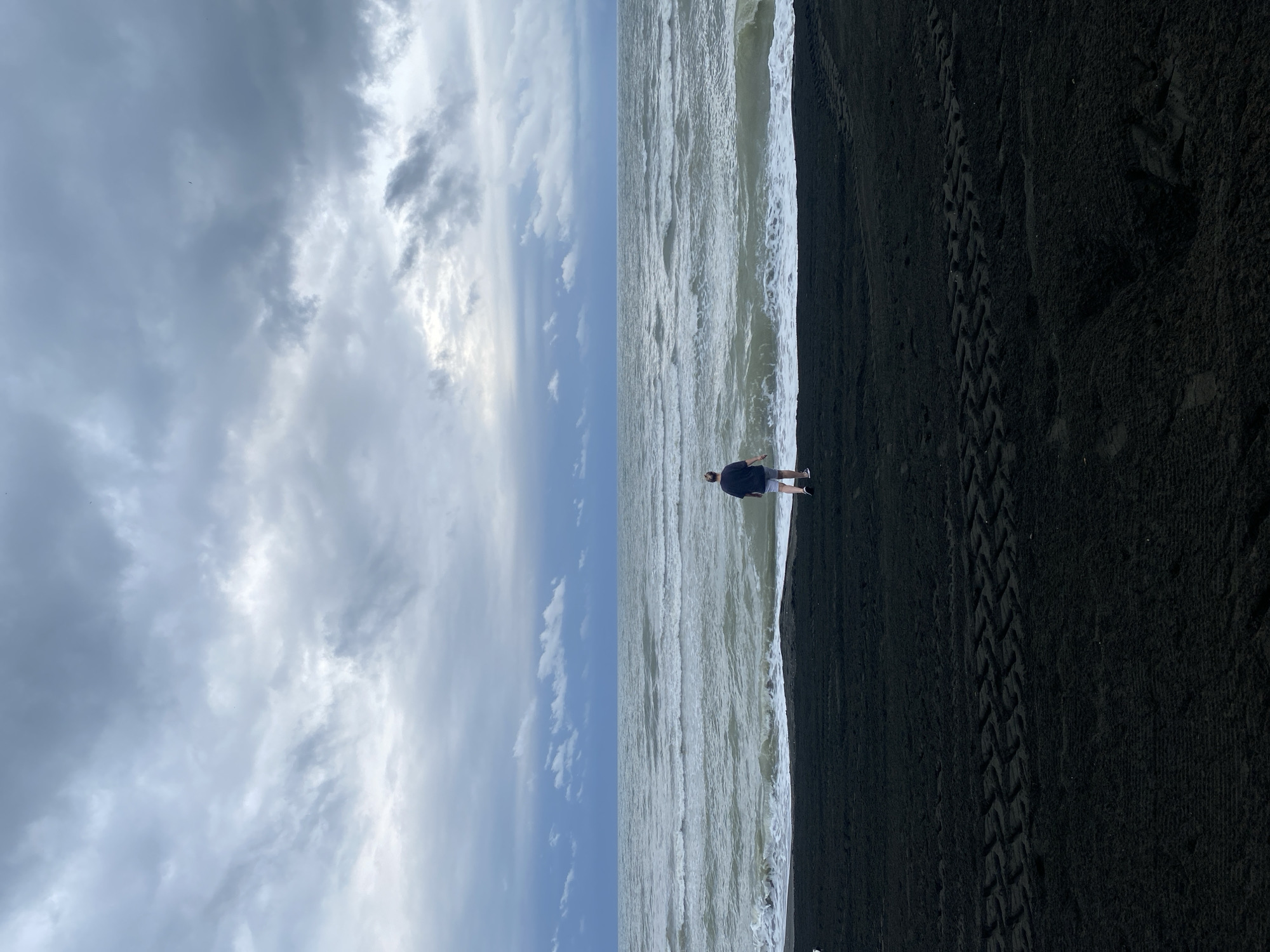 A person stands facing the sea on a black sand beach. The sky is overcast with dark, heavy clouds. The waves are choppy and white-capped. Tire tracks are visible in the dark sand. The scene appears to be the beach in Ureki, Georgia.