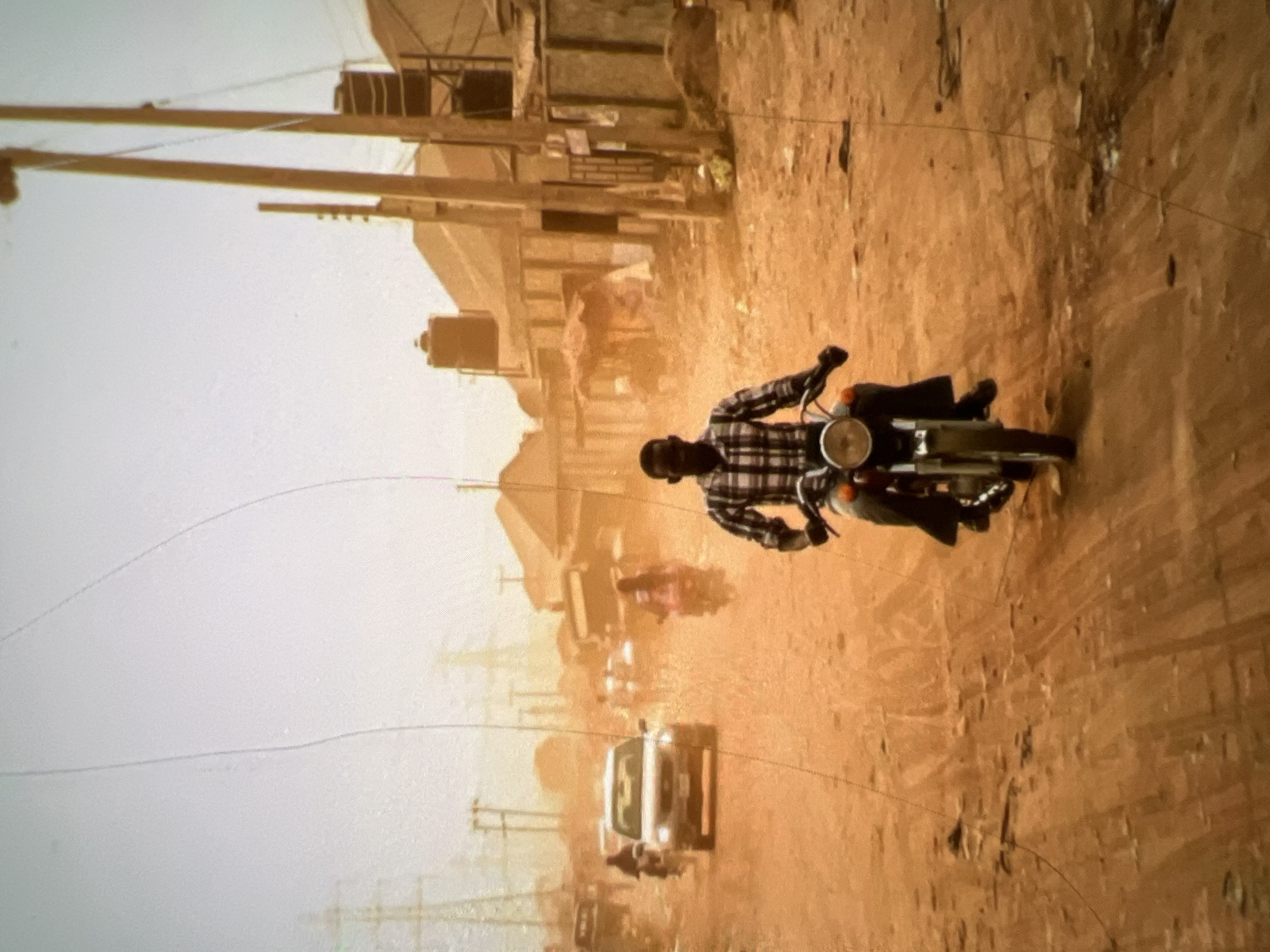 The image depicts a dusty, dirt road in what appears to be a densely populated area of Lagos, Nigeria, under hazy, overcast skies. In the foreground, a young Black man, wearing a plaid long-sleeved shirt and dark pants, is riding a motorcycle. His posture suggests he is actively riding the vehicle. In the background, other vehicles, including cars and motorcycles, are visible on the road, indicating traffic. Several buildings, some with corrugated iron roofs and others appearing to be concrete structures, line the sides of the road, with some having water tanks on top. Power lines and poles are prominent features of the urban landscape. The overall impression is one of a busy, somewhat gritty, urban environment affected by dust or poor air quality.