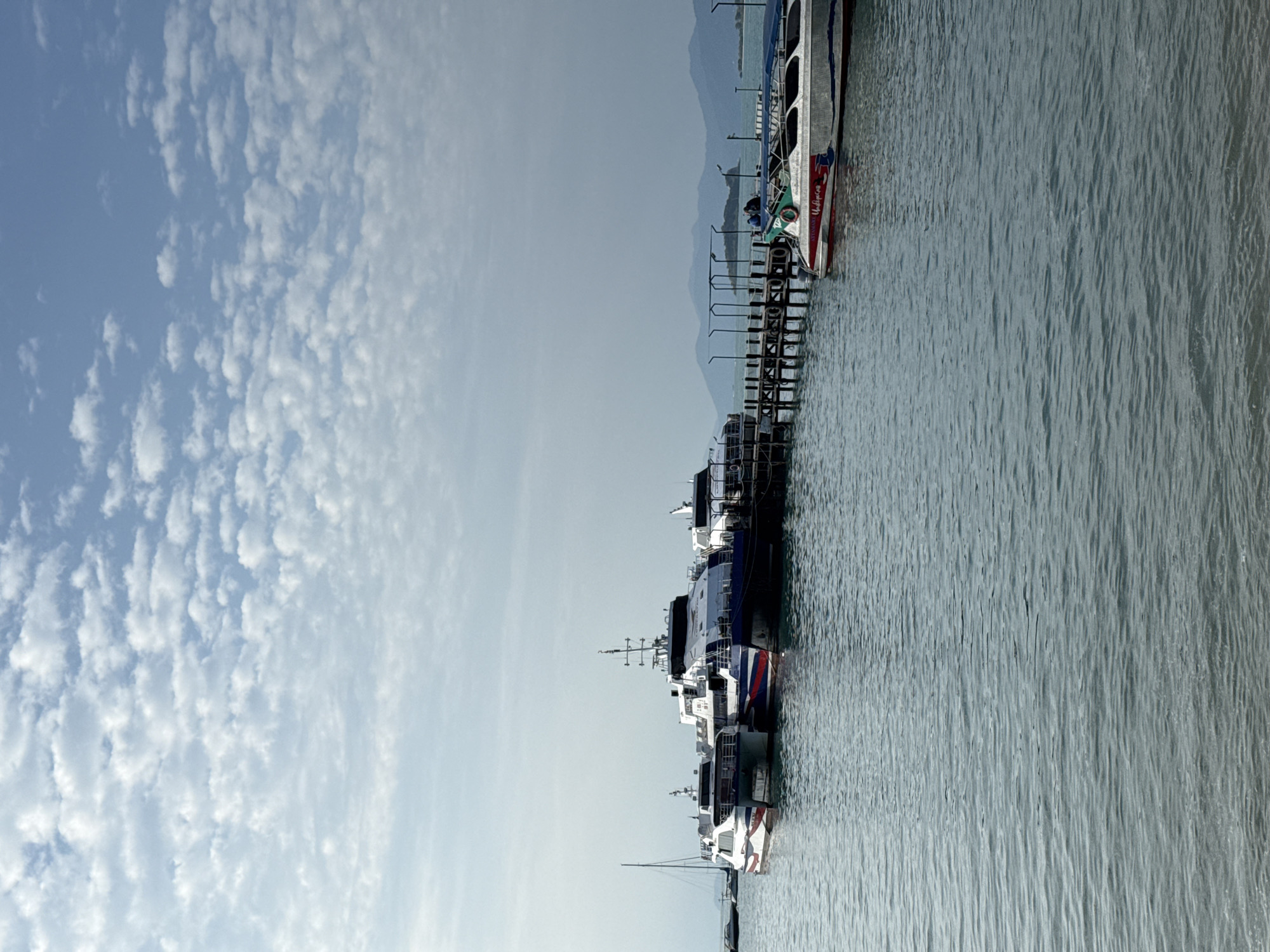 The image captures a serene scene at a pier in Ko Samui District, Thailand, under a sky dotted with cumulus clouds. Several ferry boats are docked, appearing ready for departure or arrival. One particularly large ferry, painted with blue and red accents, is prominent in the center. To its left, another ferry with red and white stripes is also moored. To the right of the central ferry, a smaller, speed boat with a white hull and red and blue trim is tied to the pier, its name visible as "STYMMANO". The pier itself is constructed of dark wood and supported by pilings. In the background, misty mountains rise against the horizon, suggesting the island's natural landscape. The water is calm, reflecting the sky and surrounding boats. No people are clearly visible in the immediate foreground, though some figures may be present on the boats or pier.