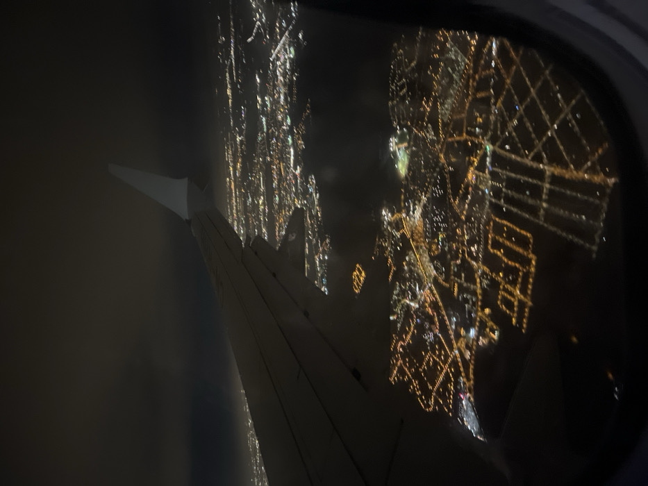 A night aerial view from an airplane window shows the wing and cityscape lights. The city lights are blurred, forming grid-like patterns and illuminated lines. Part of the plane's wing is visible in the lower left corner, casting shadows. The window frame is partially seen. Outside the window, the dark sky contrasts sharply with the bright city lights.