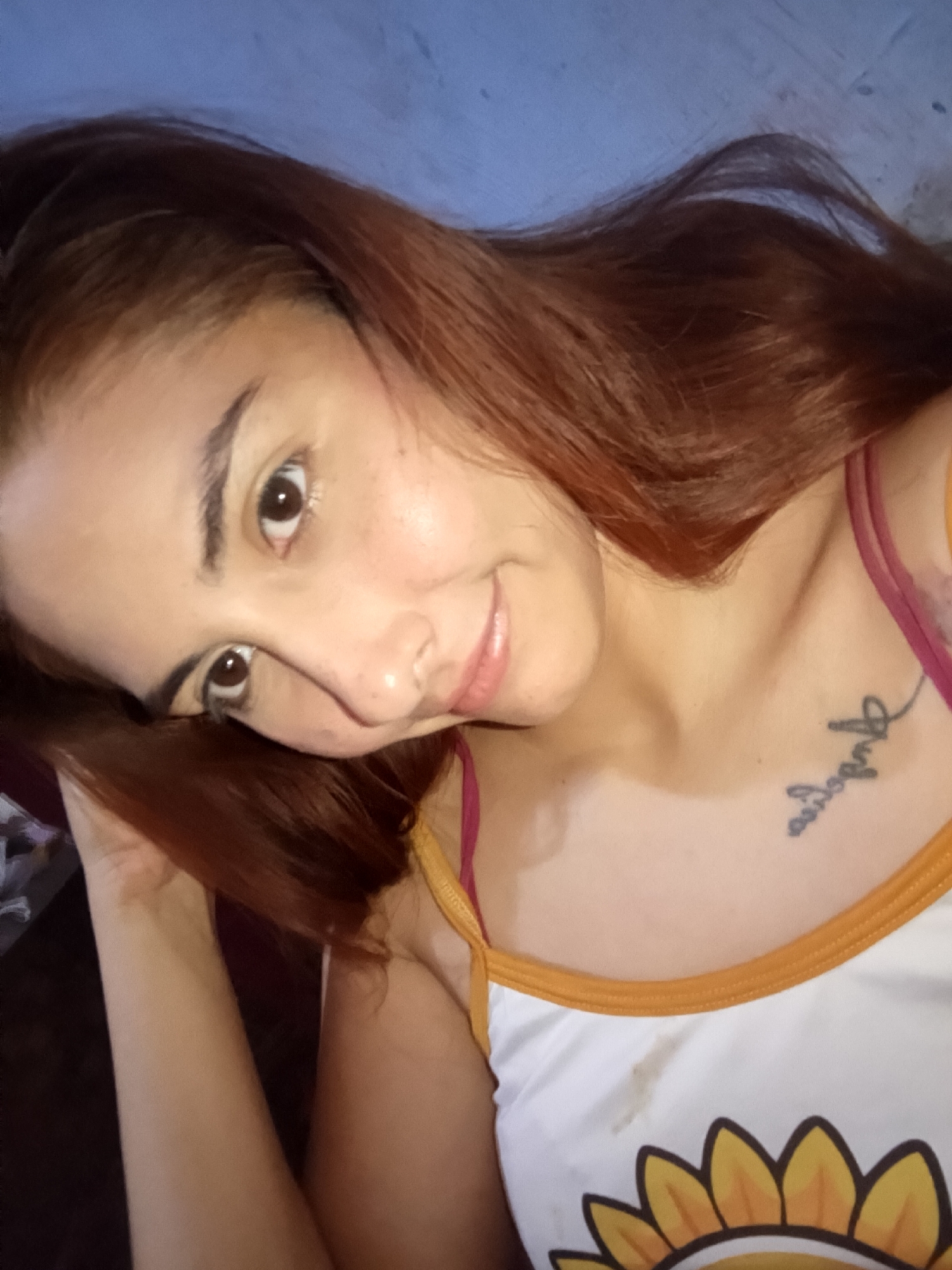 A woman with auburn hair is posing for a selfie. She is wearing a white tank top with a yellow and orange sunflower design. The tank top has orange trim and a red strap. A tattoo is visible on her upper chest. The background is a mottled blue color. There is no evidence to place this image in Juan Griego, Venezuela.