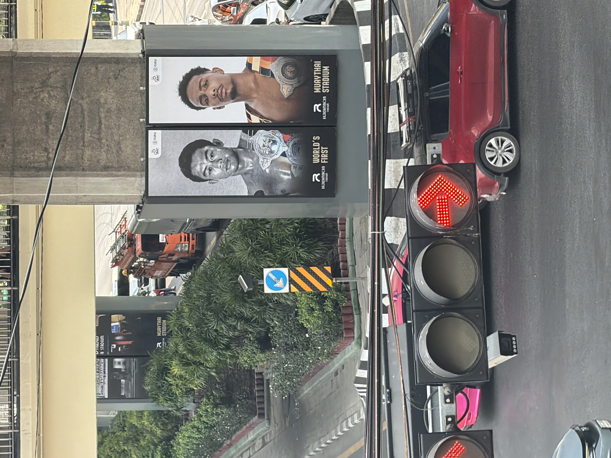 An outdoor, eye-level street scene in what appears to be Bangkok, Thailand, captured during daylight hours. The image depicts a section of a road with various urban elements.
In the foreground to the right, a vertical traffic light pole displays two illuminated red arrow signals pointing upwards, indicating that vehicles cannot proceed straight ahead. A red car is visible stopped near the traffic light on what appears to be the left side of the road, typical for Thailand. Behind the traffic light and car, a wide, dark gray road is visible.
To the left of the road, two large billboards are mounted vertically on a support structure, likely part of an elevated road or train line. The top billboard features a color portrait of a male Muaythai fighter wearing a championship belt. Text on this poster reads "MUAYTHAI STADIUM" and "RAJADAMNERN STADIUM," along with the "R" logo. The bottom billboard shows a black and white portrait of another male Muaythai fighter, also with a championship belt, with text reading "WORLD'S FIRST" and "RAJADAMNERN STADIUM." Both posters also feature the "SAT" logo and what appears to be Thai script "กฟภ."
Further left, lush green trees and bushes line the street. A blue circular road sign with a white arrow pointing diagonally downwards to the right is visible next to a yellow and black striped hazard sign. In the background, an orange utility truck with a ladder is parked, and parts of buildings and what looks like an elevated concrete structure are visible. Electrical wires crisscross the upper part of the frame. The overall setting is a busy city environment with prominent advertising for a Muaythai venue.