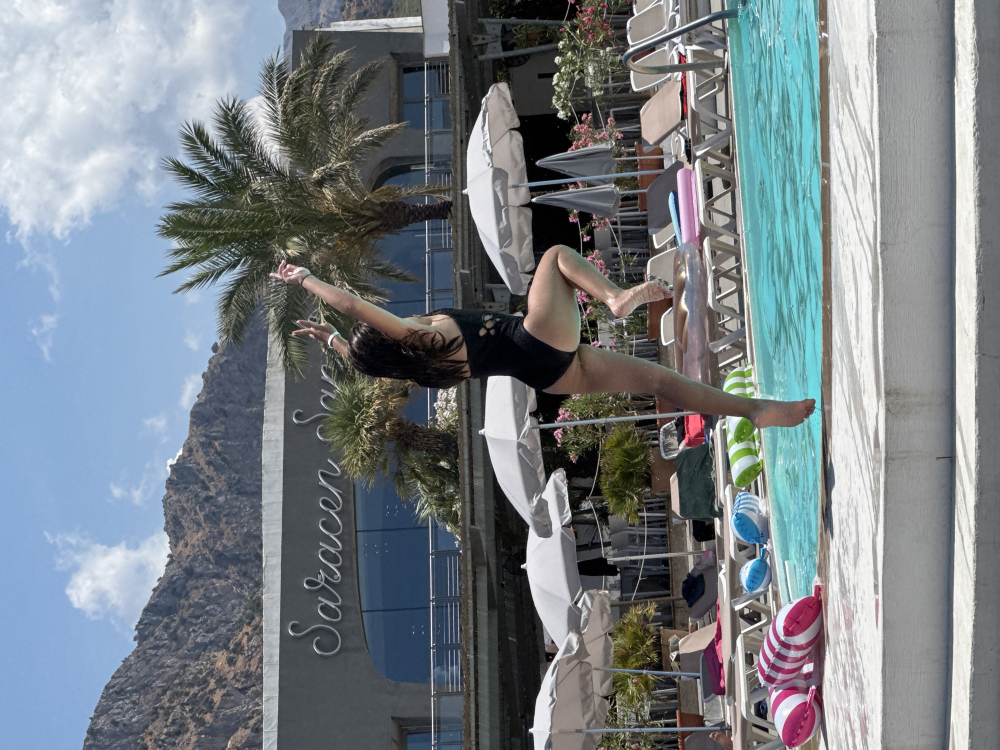 A young woman in a black one-piece swimsuit is captured mid-air, leaping into a turquoise swimming pool. Her body is positioned in a dynamic pose, with her arms extended upwards and her legs bent. She is located at a resort in Isola delle Femmine, Italy, evidenced by the "Saracen Sands" signage visible on the building in the background. The resort features palm trees, white umbrellas, and lounge chairs scattered around the pool area. Several inflatable pool toys, including striped pink and white, blue, and green and white balls, are floating on the water or near the pool's edge. The scene is set against a backdrop of a rocky mountain and a bright blue sky with scattered clouds.